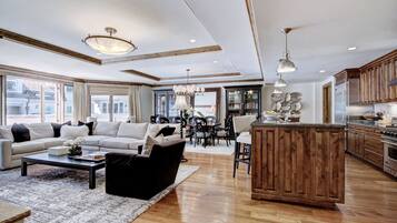 A spacious living room with a large sofa, a coffee table, and a dining area with chairs and a table. There is a kitchen area with wooden cabinets and a refrigerator.