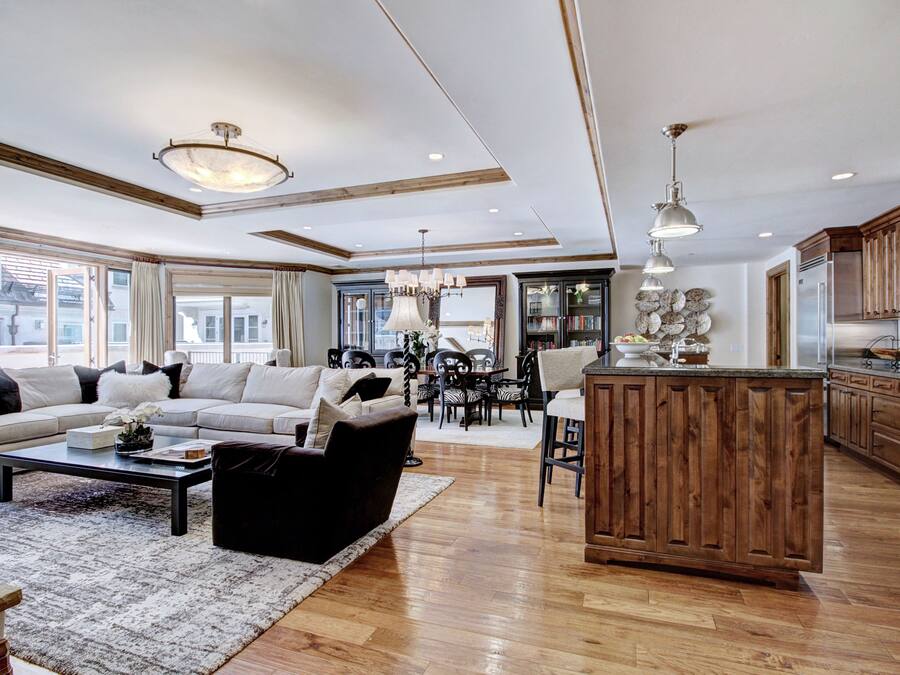 A spacious living room with a large sofa, a coffee table, and a dining area with chairs and a table. There is a kitchen area with wooden cabinets and a refrigerator.