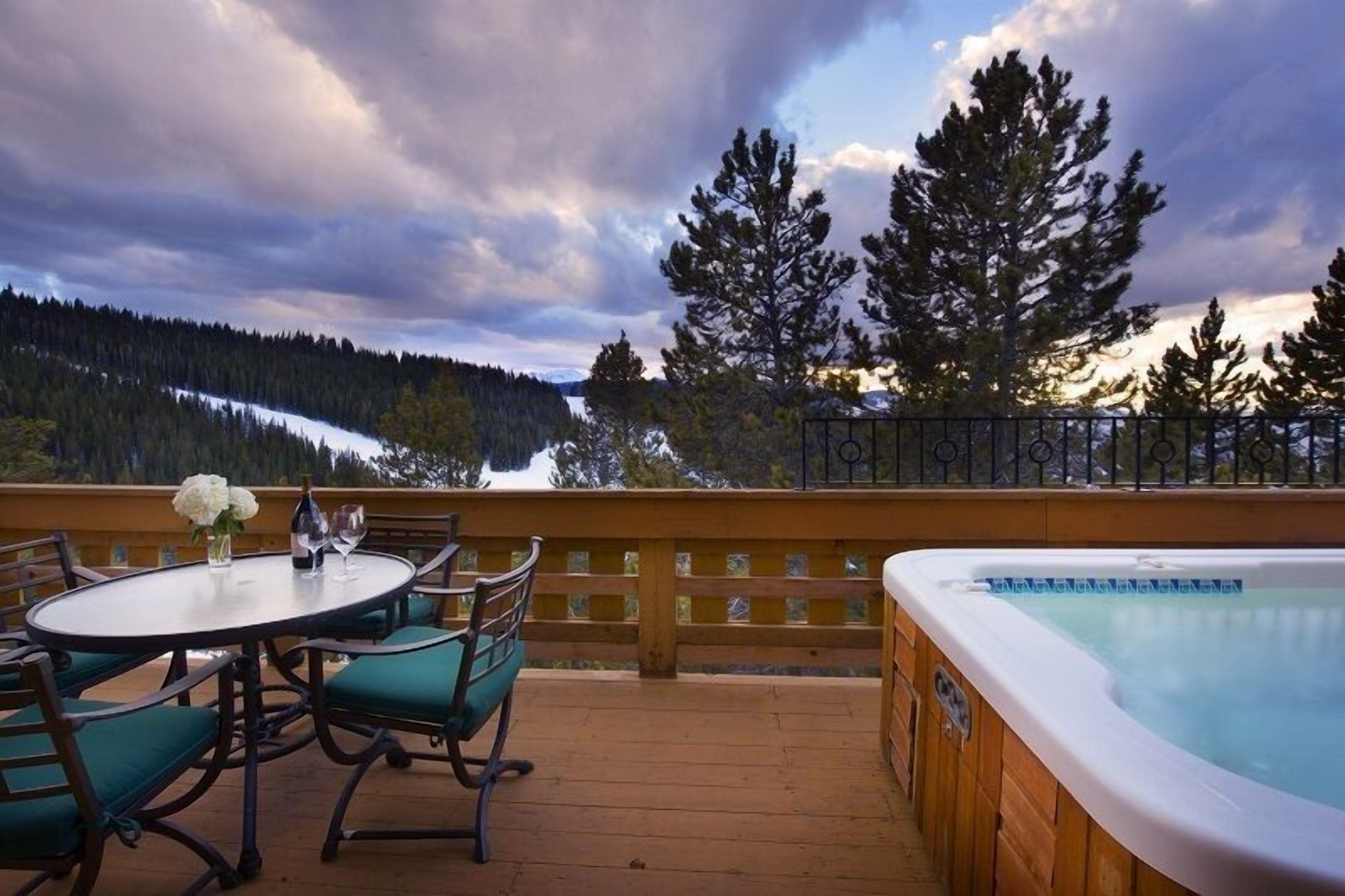 A wooden deck with a hot tub and a table with chairs, overlooking a snowy mountain range.
