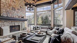 A cozy living room with a stone fireplace, large windows offering a forest view, and modern furnishings. - The Arrabelle, A RockResort, by Vail Resorts (Vail)