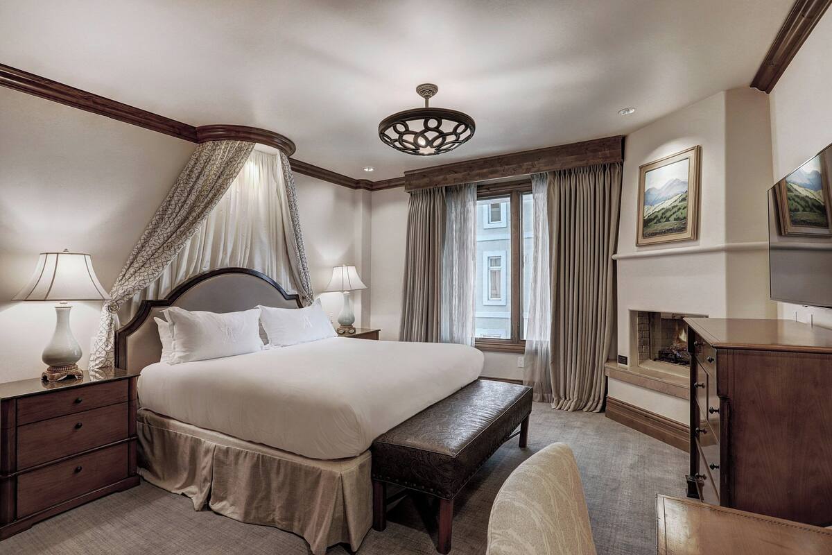 A hotel room with a large bed, a bench, a nightstand, a lamp, a fireplace, and a television.