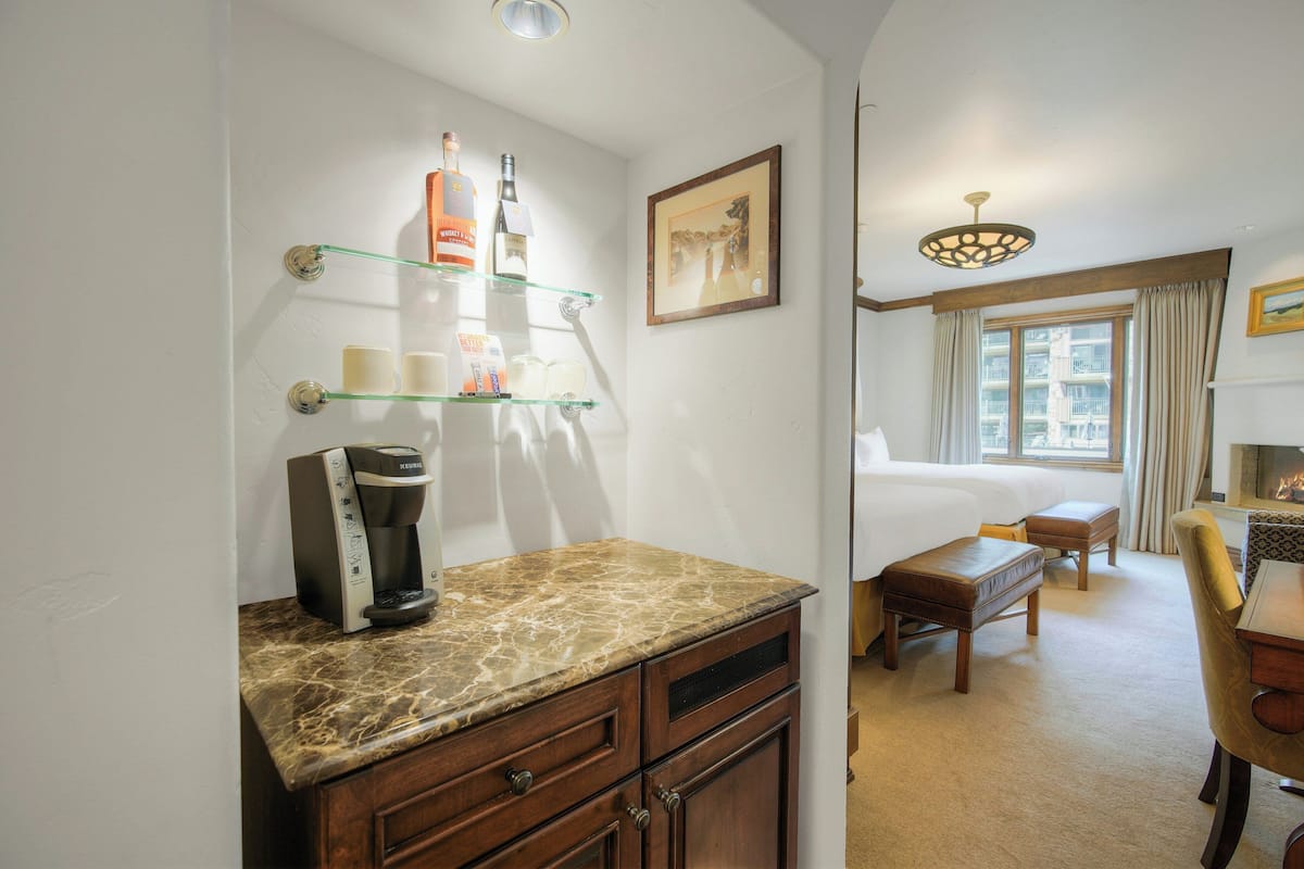 A hotel room with a bed, a coffee maker, a desk with a chair, a window with curtains, and a painting on the wall.