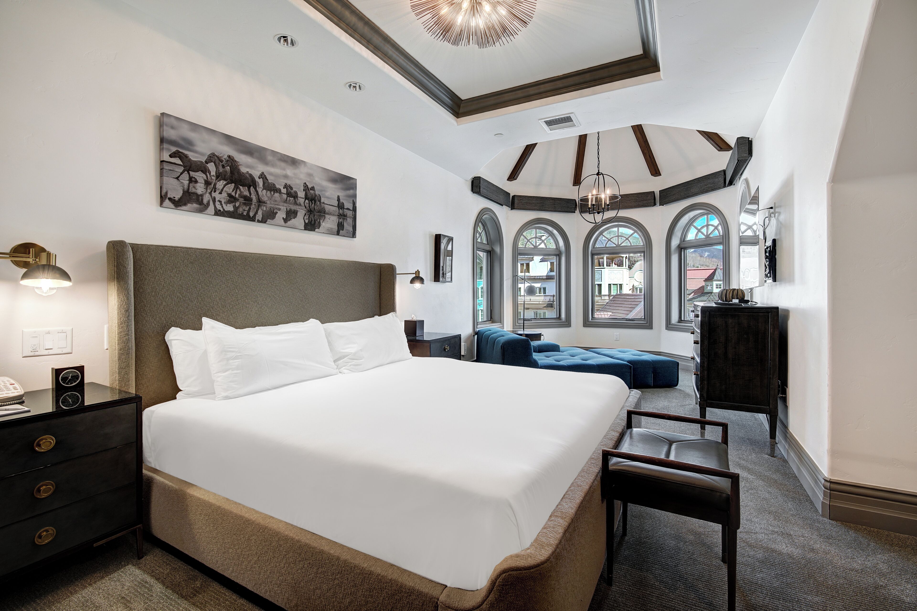 A spacious bedroom with a large bed, a desk, and a chair. There are arched windows and a chandelier.