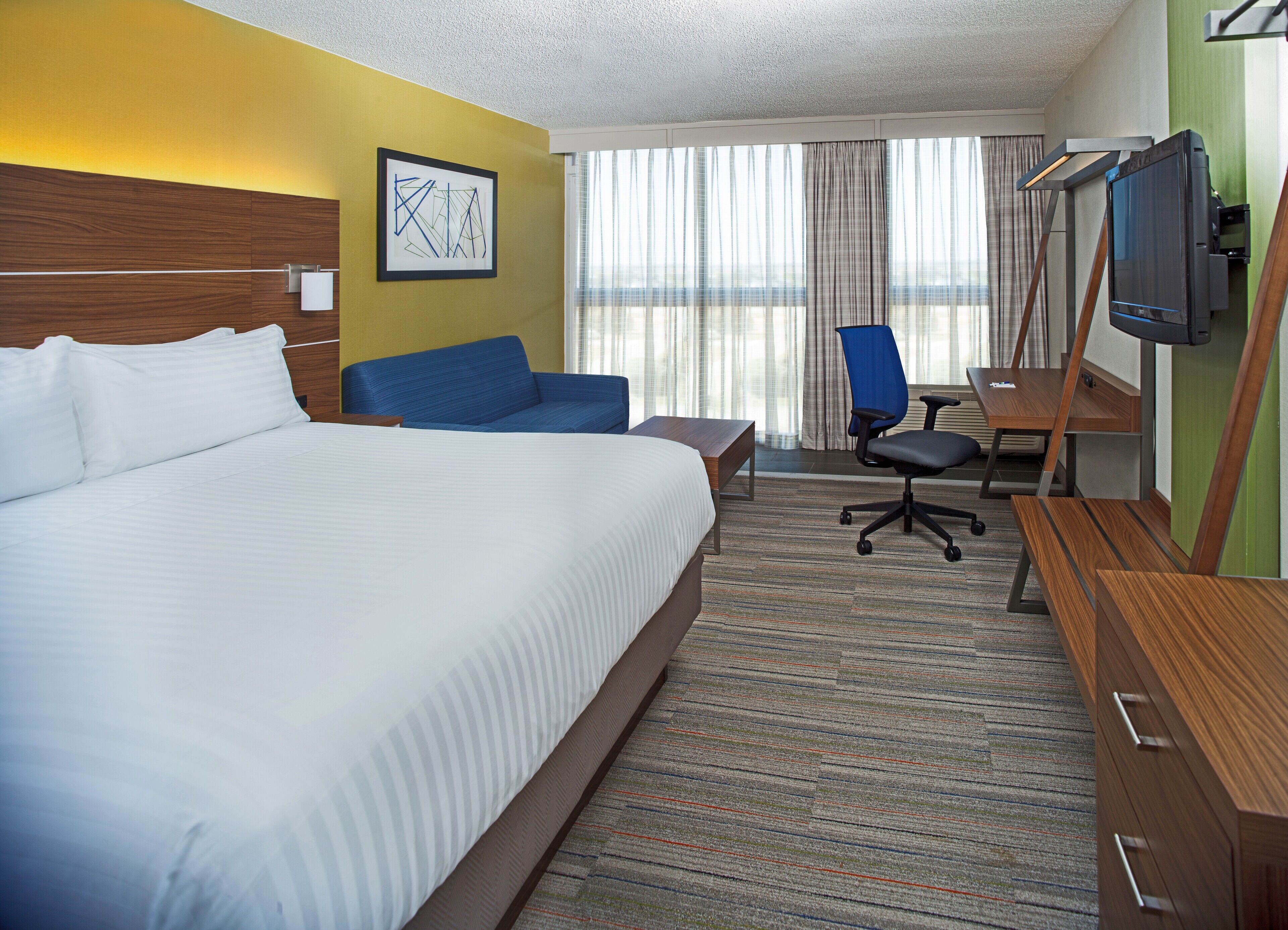 A hotel room with a large bed, a blue armchair, a desk with a chair, a flat-screen TV, and a window with curtains.