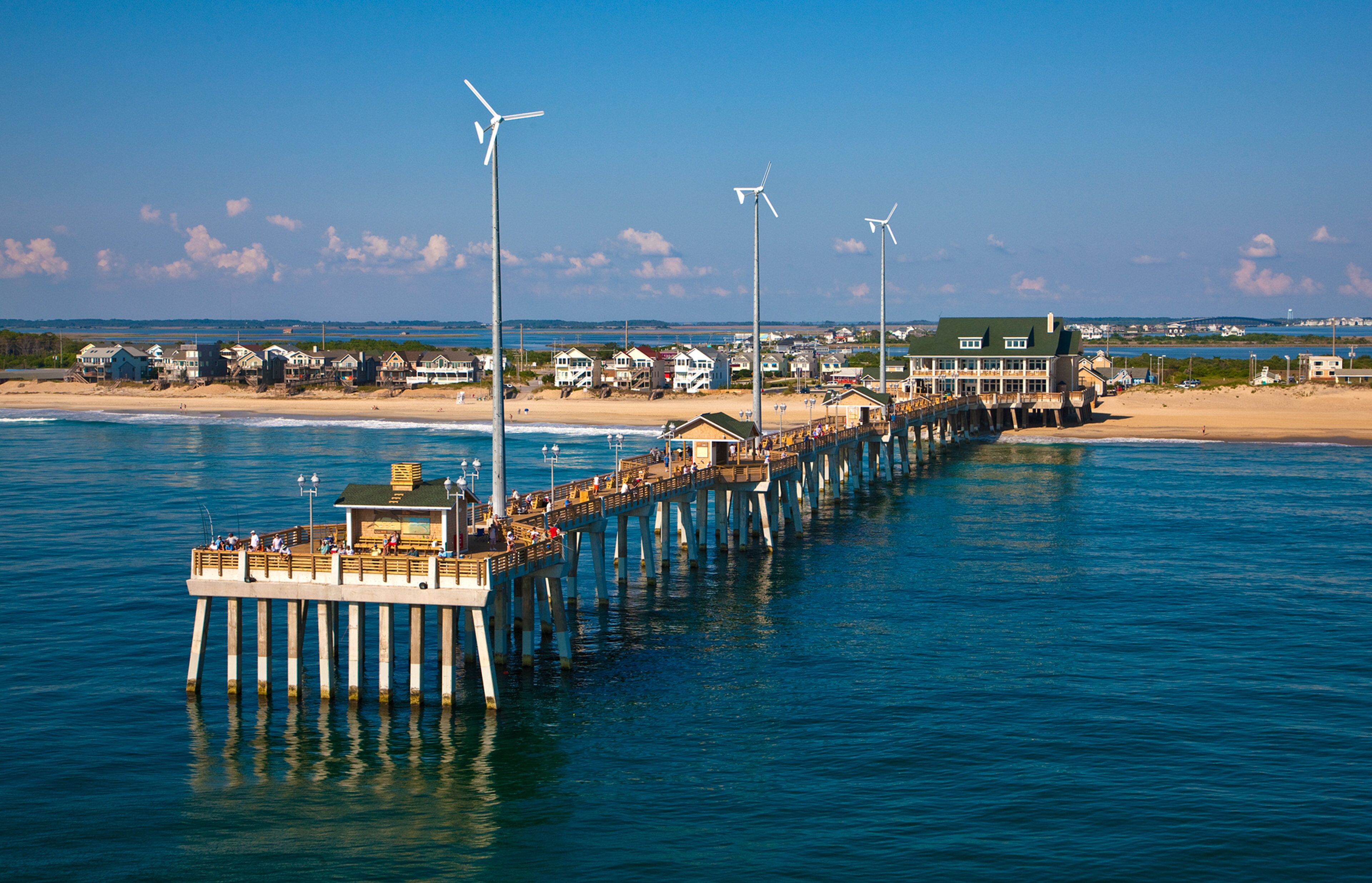 A pier extending into the water with buildings and a beach in the background.