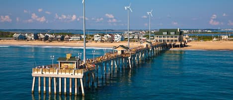 Un muelle que se adentra en el agua, con edificios y una playa al fondo.