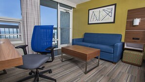 A hotel room with a blue sofa, a wooden coffee table, a blue office chair, a patterned ottoman, and a large window offering a view of the ocean. - Holiday Inn Express Nags Head Oceanfront by IHG (Nags Head)