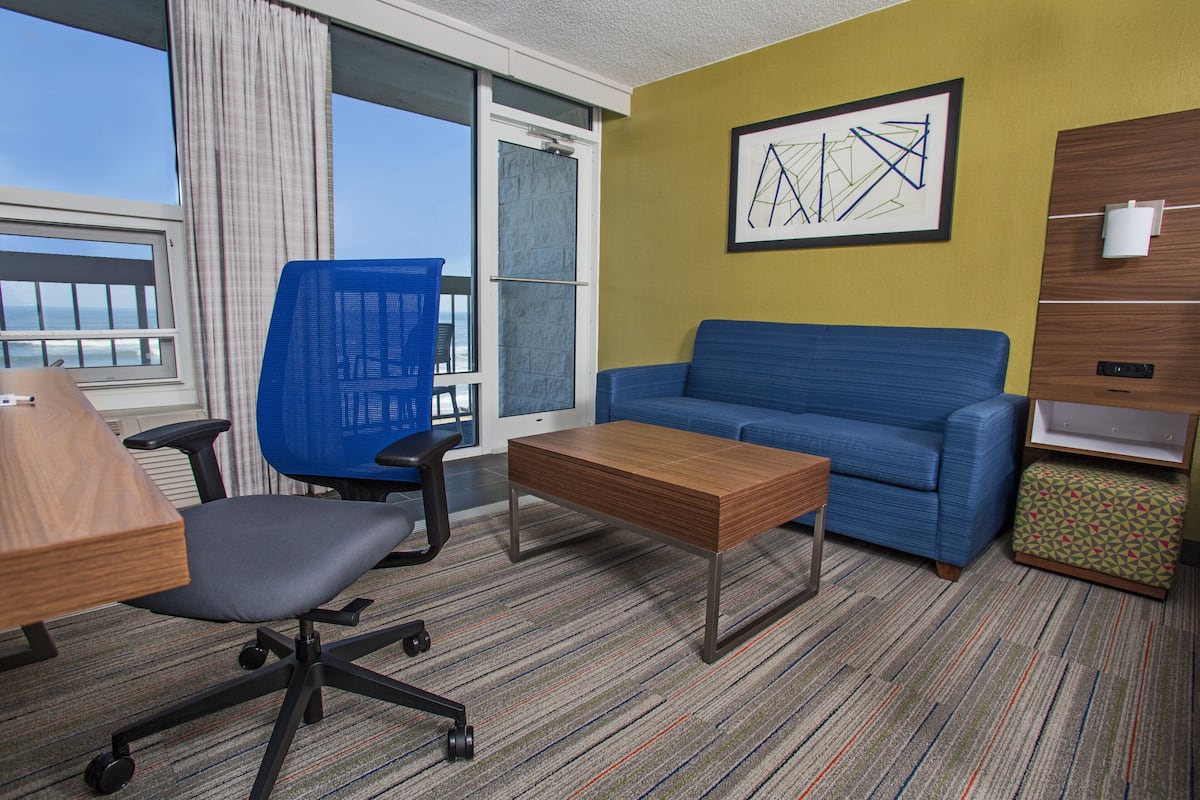 Une chambre d’hôtel avec un canapé bleu, une table basse en bois, un fauteuil de bureau bleu, un pouf à motifs et une grande fenêtre donnant sur l’océan.