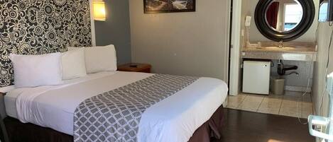 Una habitación de hotel con cama, pared con estampado, pintura de un faro, espejo redondo y una pequeña kitchenette.