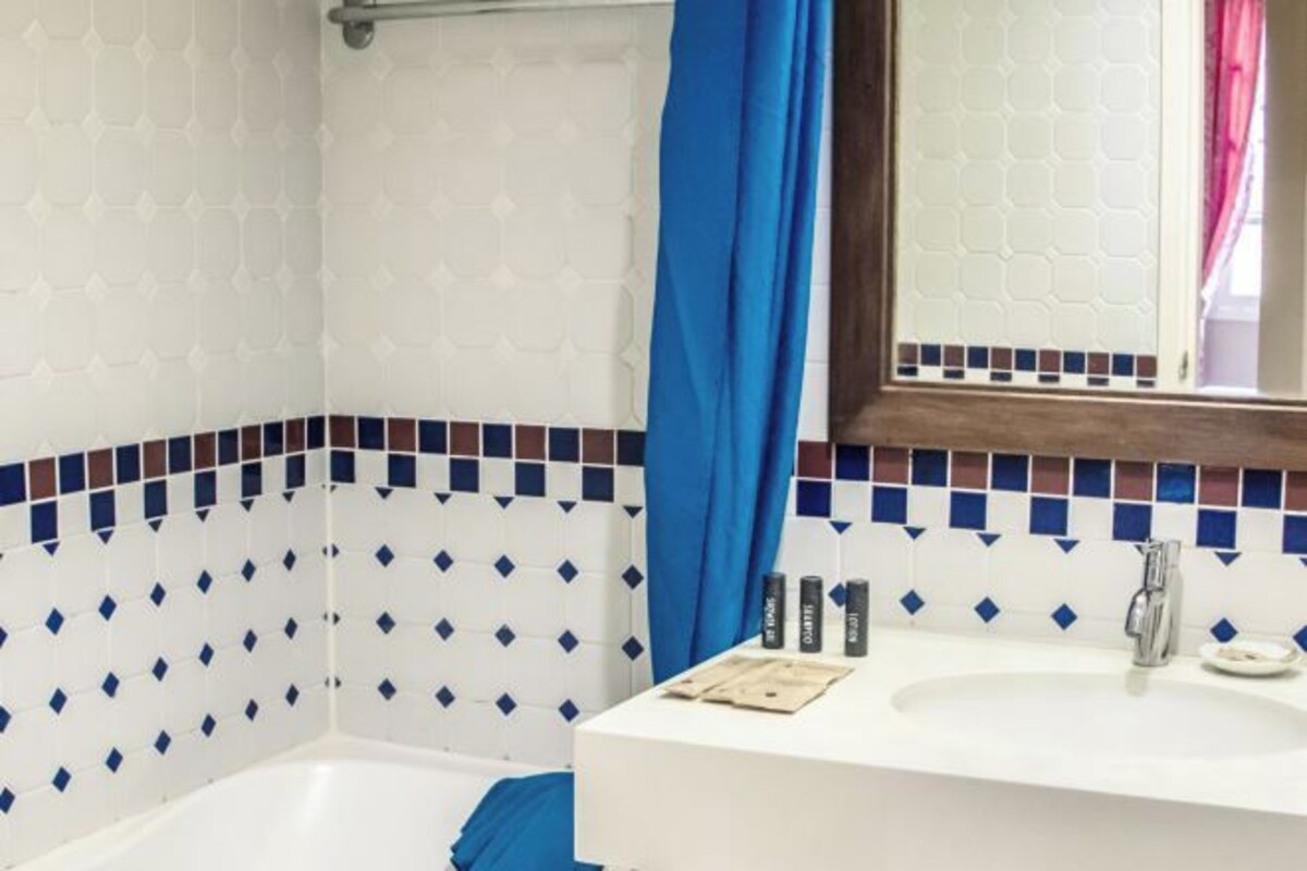 Un baño con un lavabo blanco, azulejos blancos y azules con diseño, una cortina de ducha azul y un espejo con marco de madera.