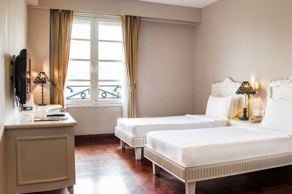 A hotel room with two single beds, a desk with a computer and lamp, and a window with curtains.