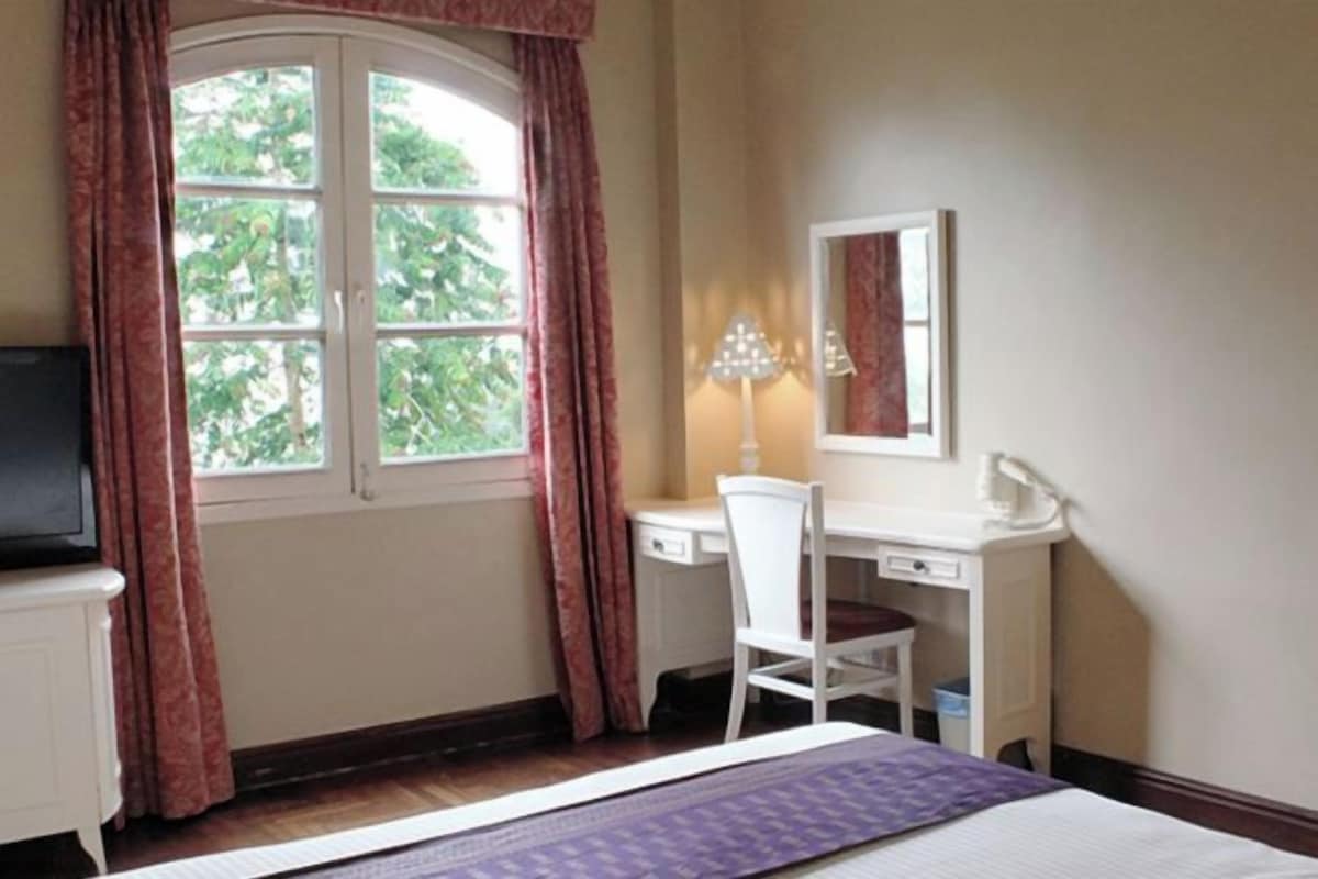 A bedroom with a bed, a desk, a chair, a lamp, and a window with curtains.