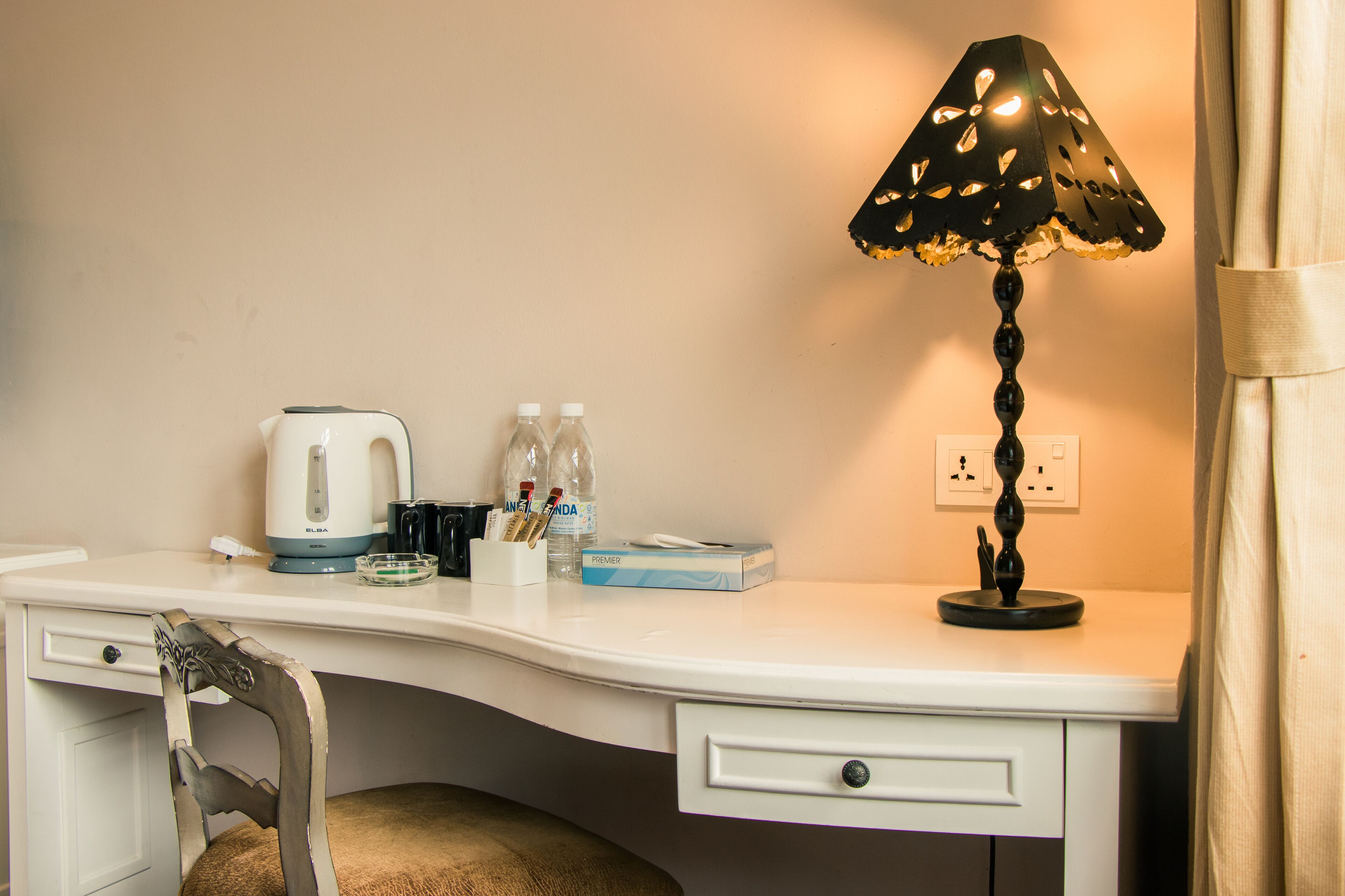 A white desk with a black lamp, a kettle, two bottles, a box of tissues, and a chair.
