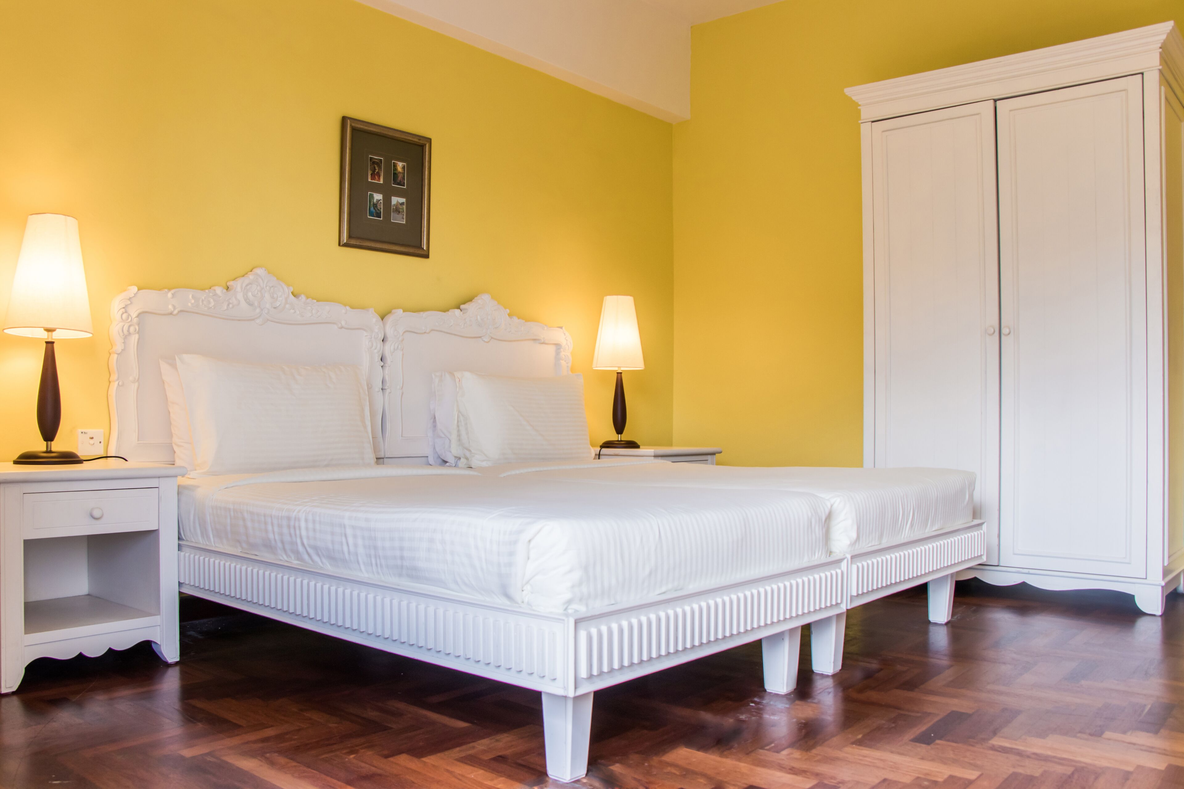 Een slaapkamer met een wit bed, twee bedlampen, een nachtkastje, een kledingkast en een ingelijste foto aan de muur.