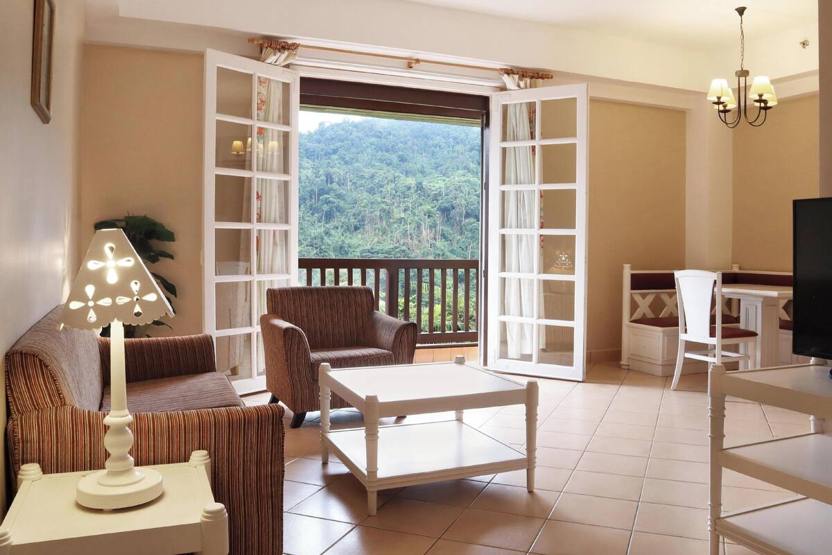 A cozy living room with a sofa, armchair, and coffee table, a balcony with a scenic view, and a television on a stand.