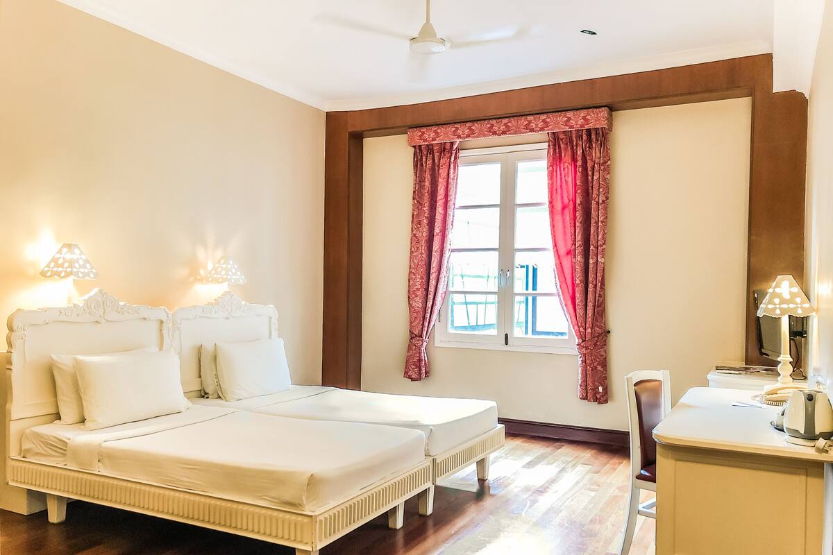A double bed with a wooden frame, a bedside table with a lamp, and a window with red curtains.