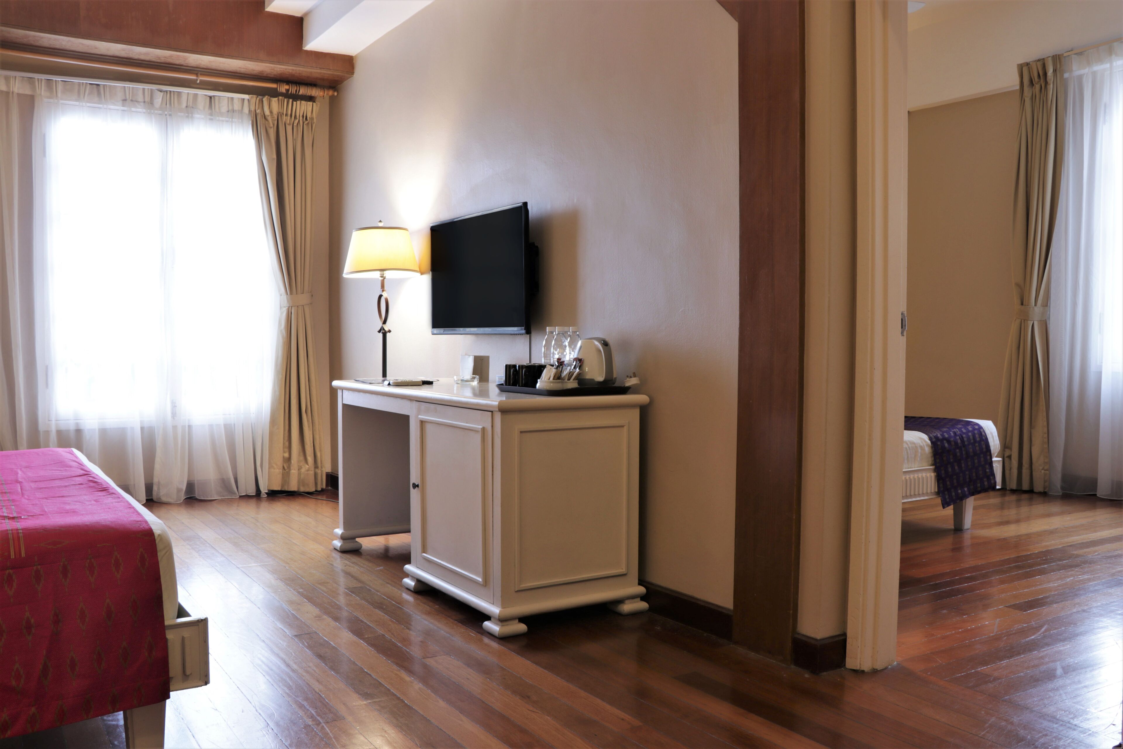 A hotel room with a wooden floor, a bed with a red cover, a desk with a lamp, a television, and a bathroom visible through an open door.