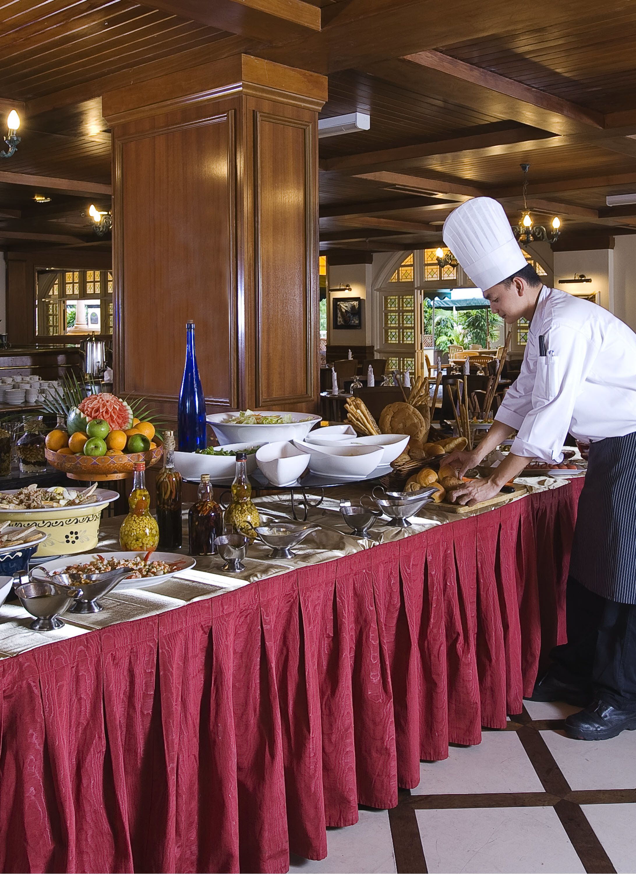 A chef preparing food in a well-lit, spacious kitchen with wooden beams on the ceiling and a large buffet table covered with a red cloth.