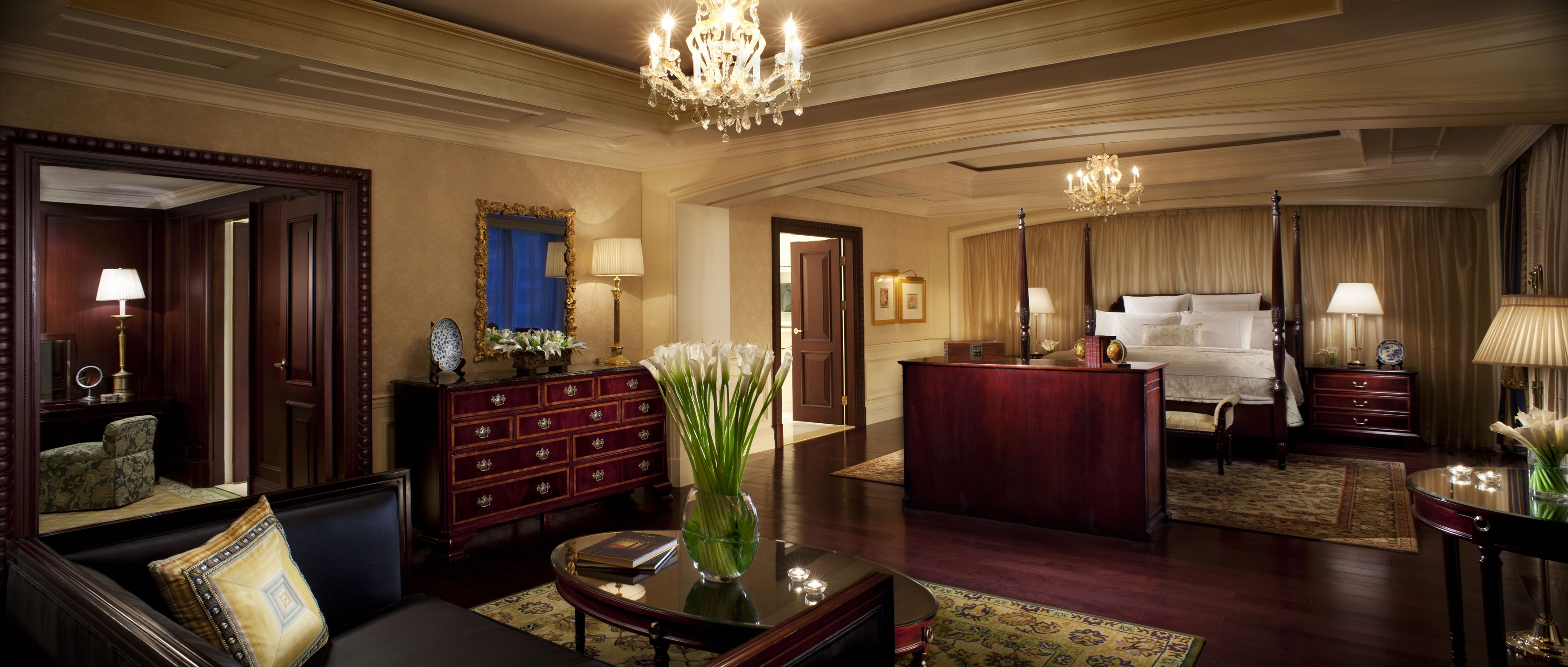 A luxurious hotel room with a large bed, a sitting area with a sofa and a coffee table, a desk, and a chandelier.
