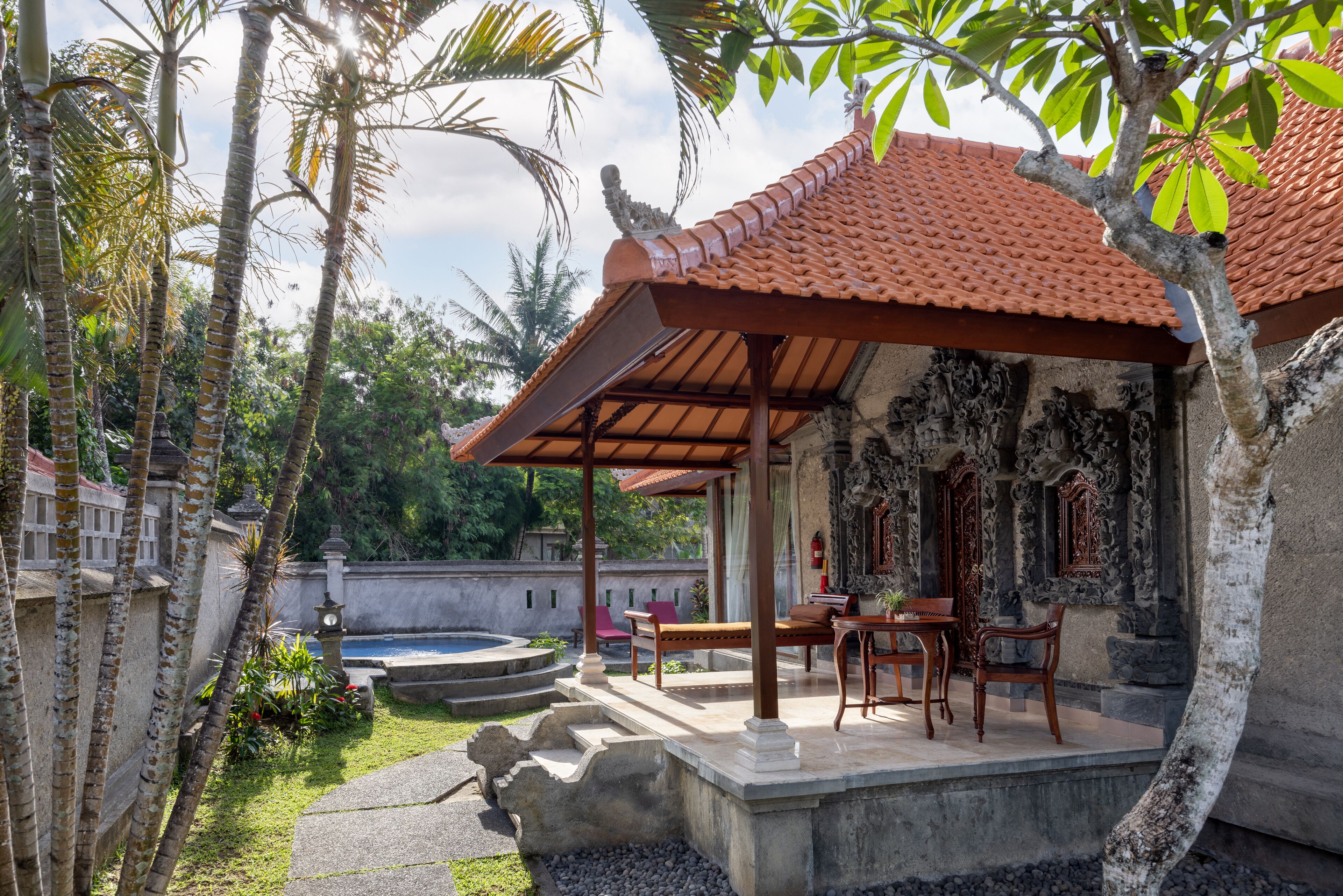 A covered outdoor seating area with a pool, surrounded by palm trees and a stone wall.