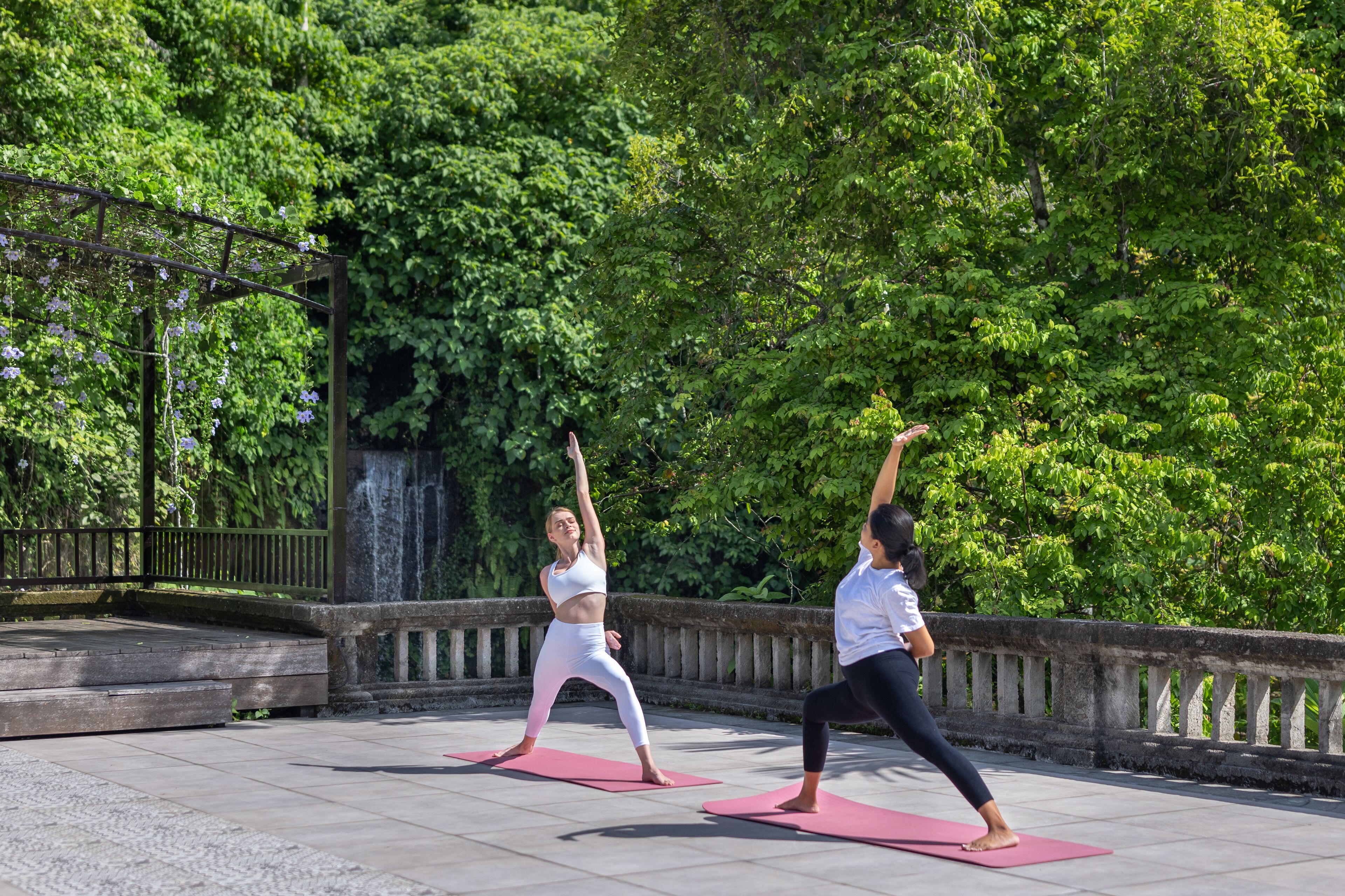 Duas pessoas praticando yoga em tapetes cor-de-rosa ao ar livre, com um parapeito de pedra e vegetação exuberante ao fundo.