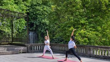 Duas pessoas praticando yoga em tapetes cor-de-rosa ao ar livre, com um parapeito de pedra e vegetação exuberante ao fundo.
