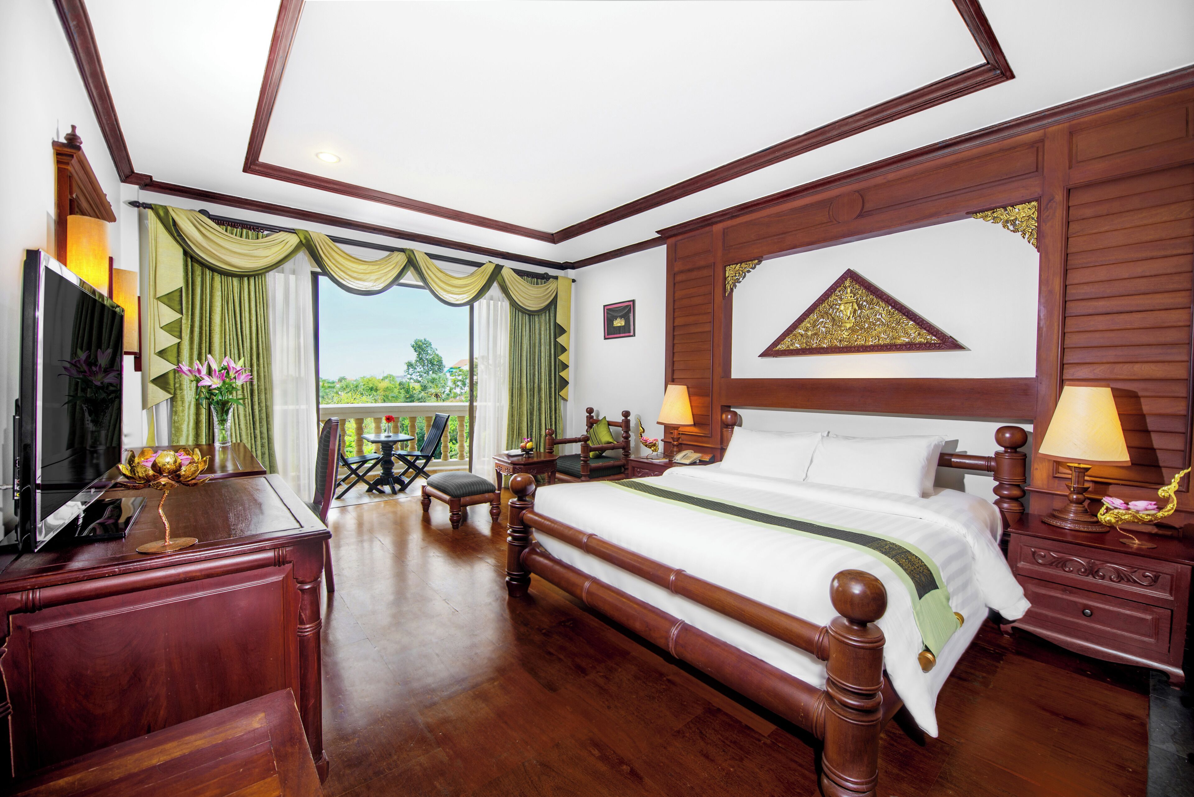 A spacious bedroom with a large bed, wooden furniture, a desk with a TV, and a balcony with a view.