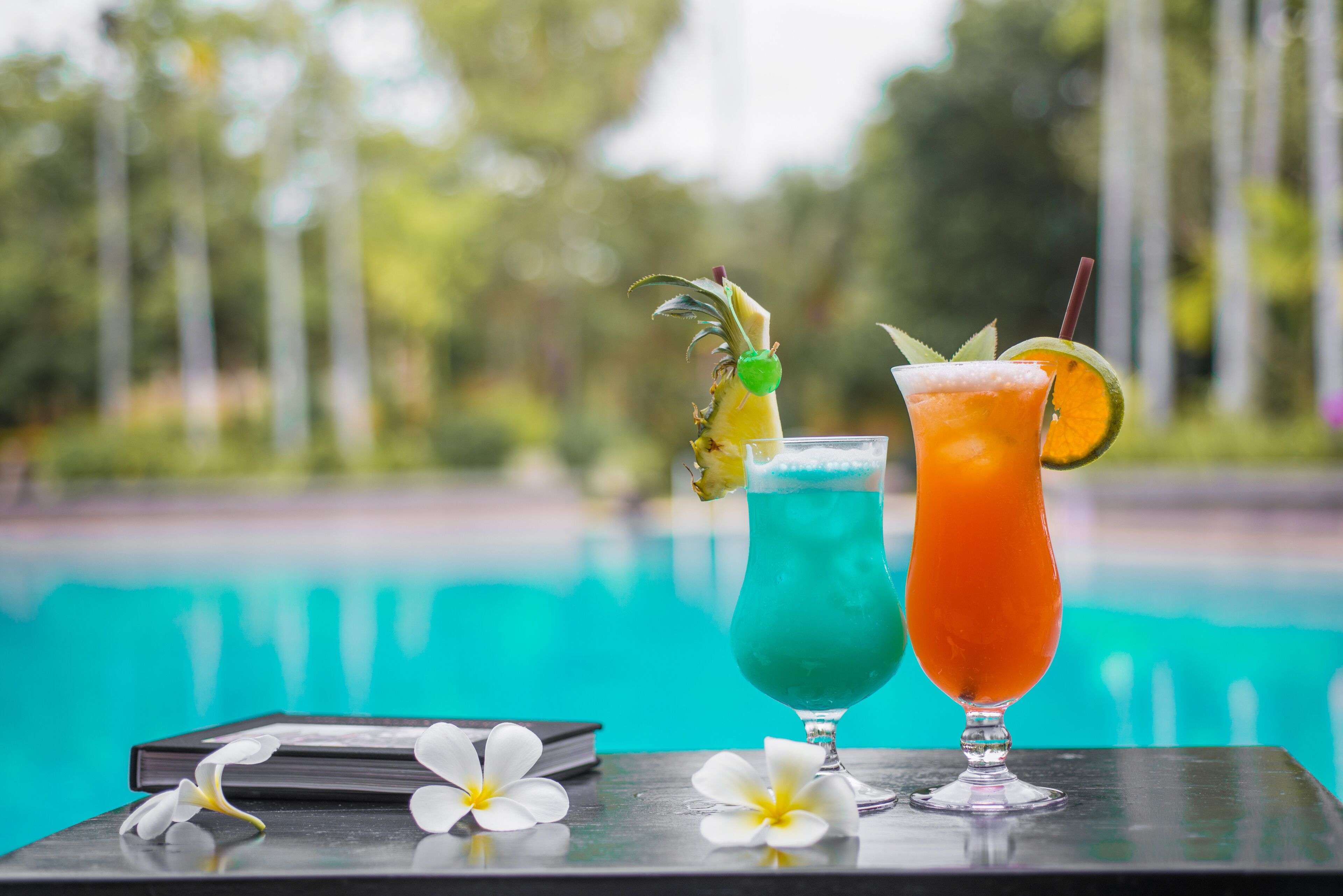 Two cocktails on a table by a pool with a blurred green background.