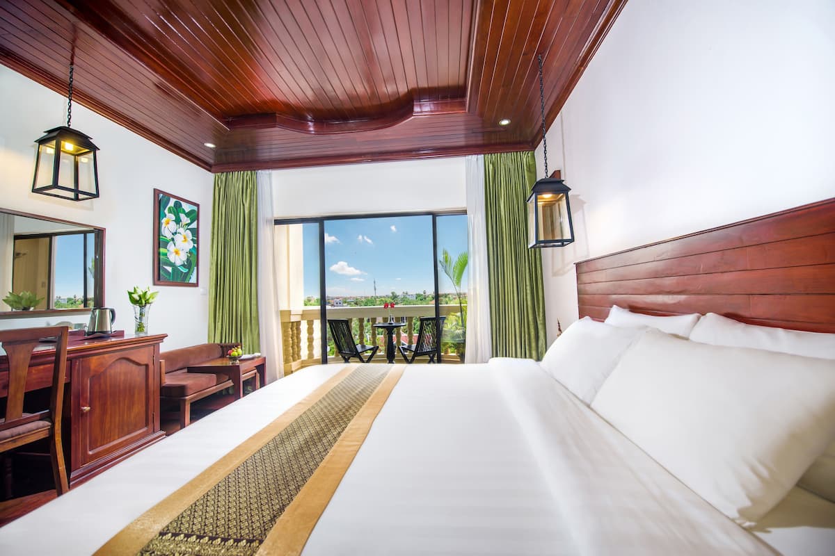 Une chambre d’hôtel avec un grand lit, une tête de lit en bois, un bureau avec des chaises, une petite table et un balcon avec vue.