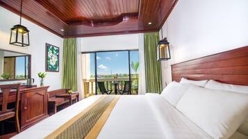 Une chambre d’hôtel avec un grand lit, une tête de lit en bois, un bureau avec des chaises, une petite table et un balcon avec vue.