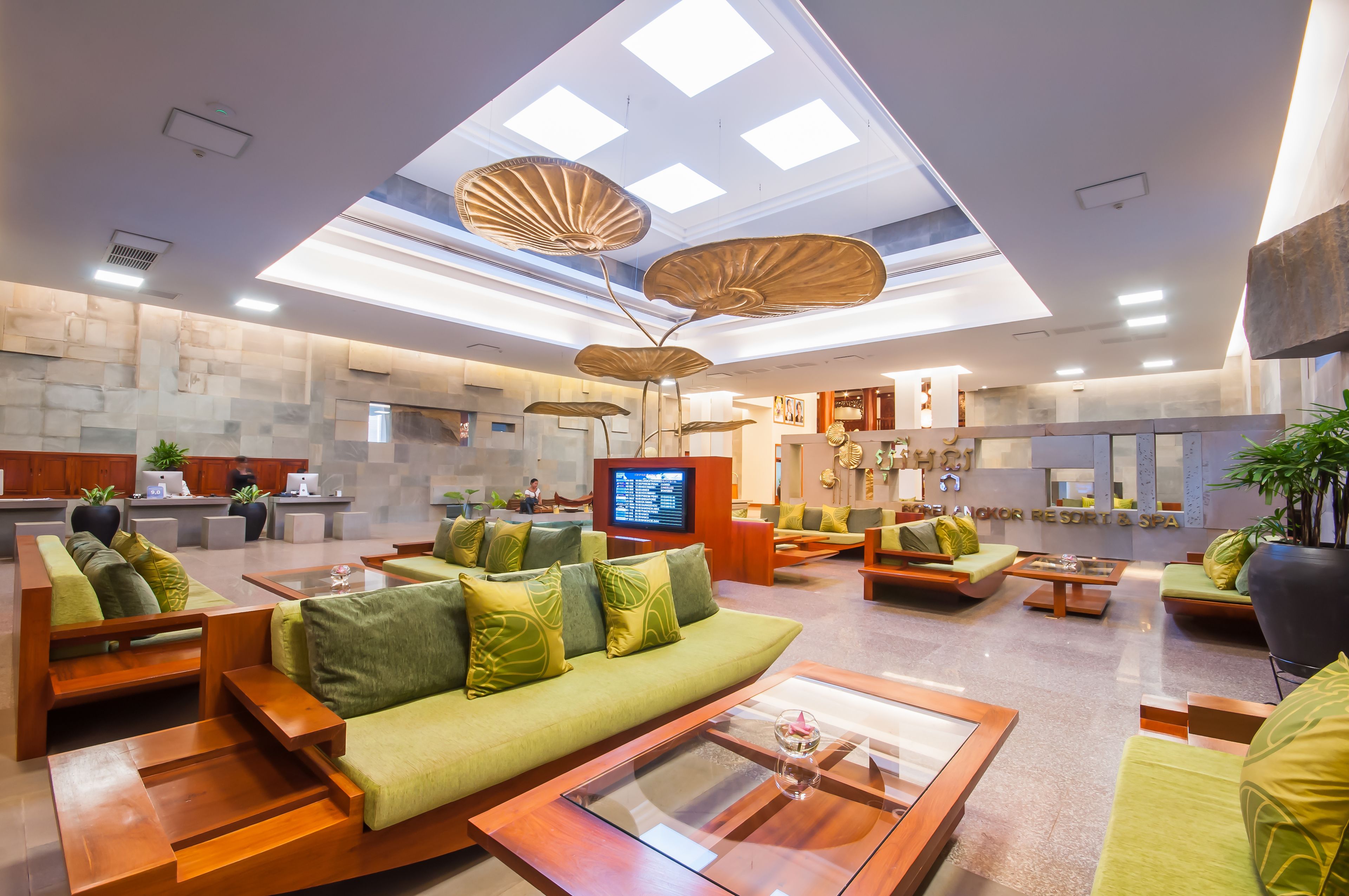 A modern hotel lobby with green sofas, wooden tables, and a large TV screen.