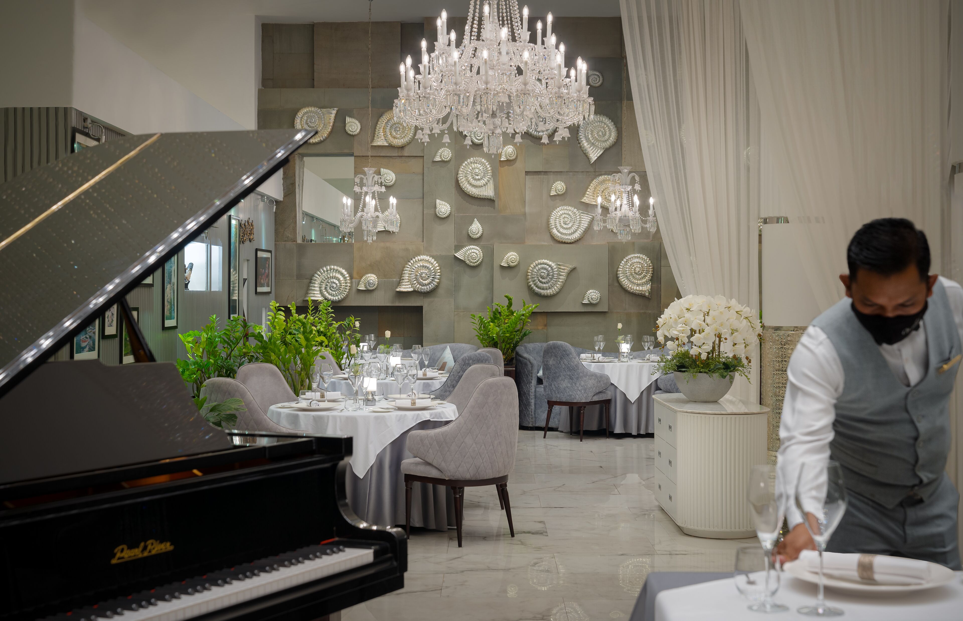 Een vleugelpiano, elegante eettafels en een ober die aan een tafel serveert in een stijlvol interieur.
