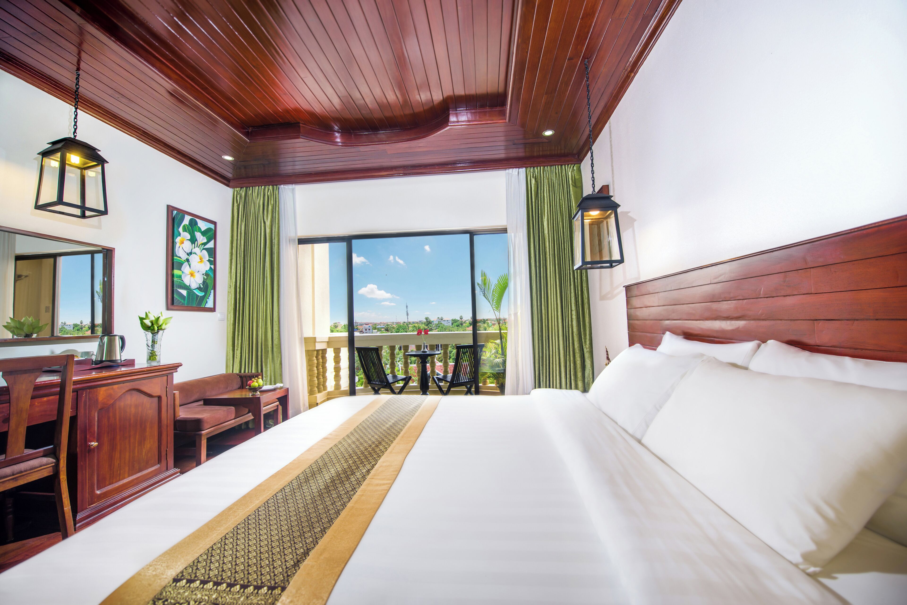 A hotel room with a large bed, wooden headboard, a desk with a chair, a small table with chairs, and a balcony with a view.
