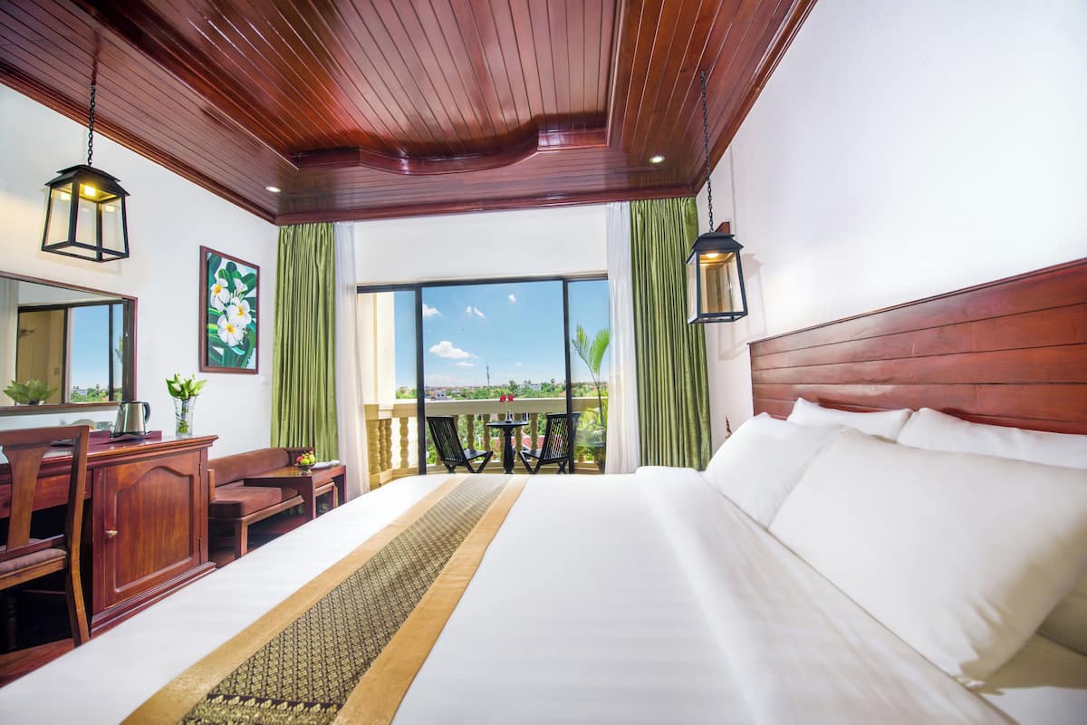 A hotel room with a large bed, wooden headboard, a desk with a chair, a small table with chairs, and a balcony with a view.