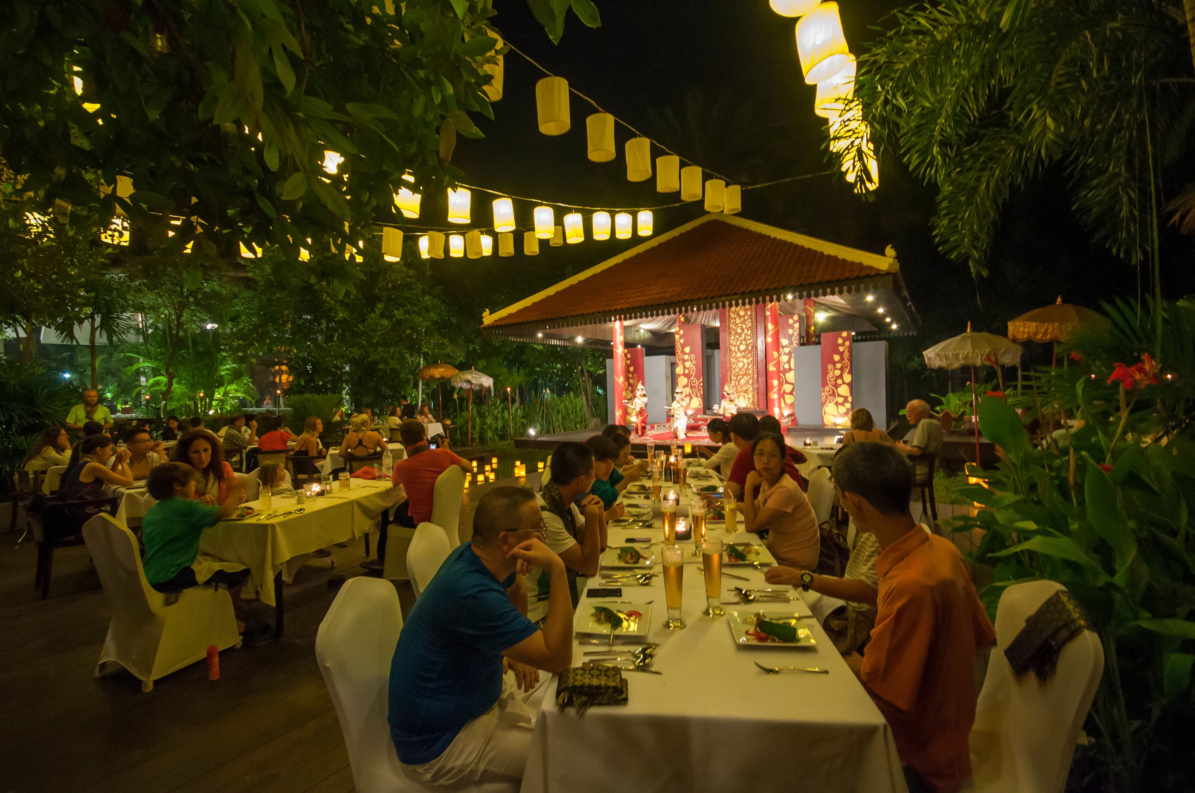 An outdoor dining area with tables set for guests, surrounded by greenery and string lights.