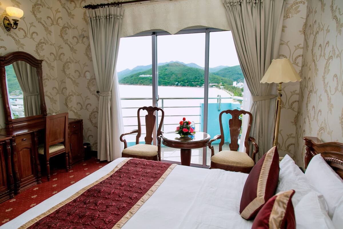 A hotel room with a large bed, a desk, two chairs, a small table with a flower vase, and a view of a body of water and mountains through large windows.