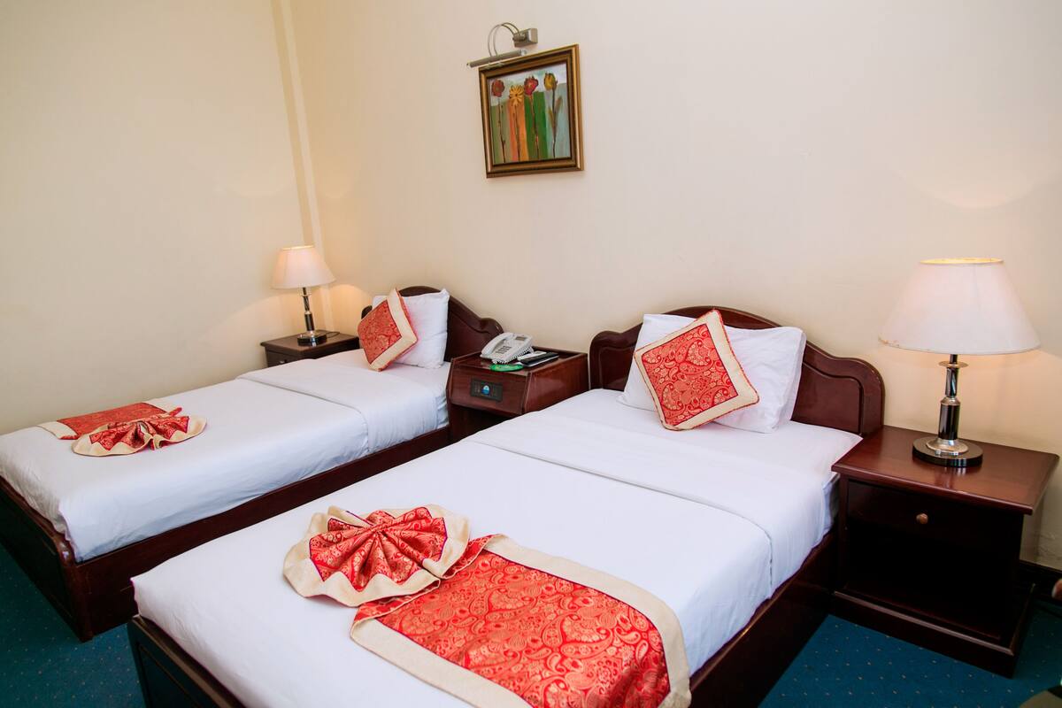 Two double beds with white linens and red decorative pillows, a bedside table with a lamp, and a wall-mounted painting.