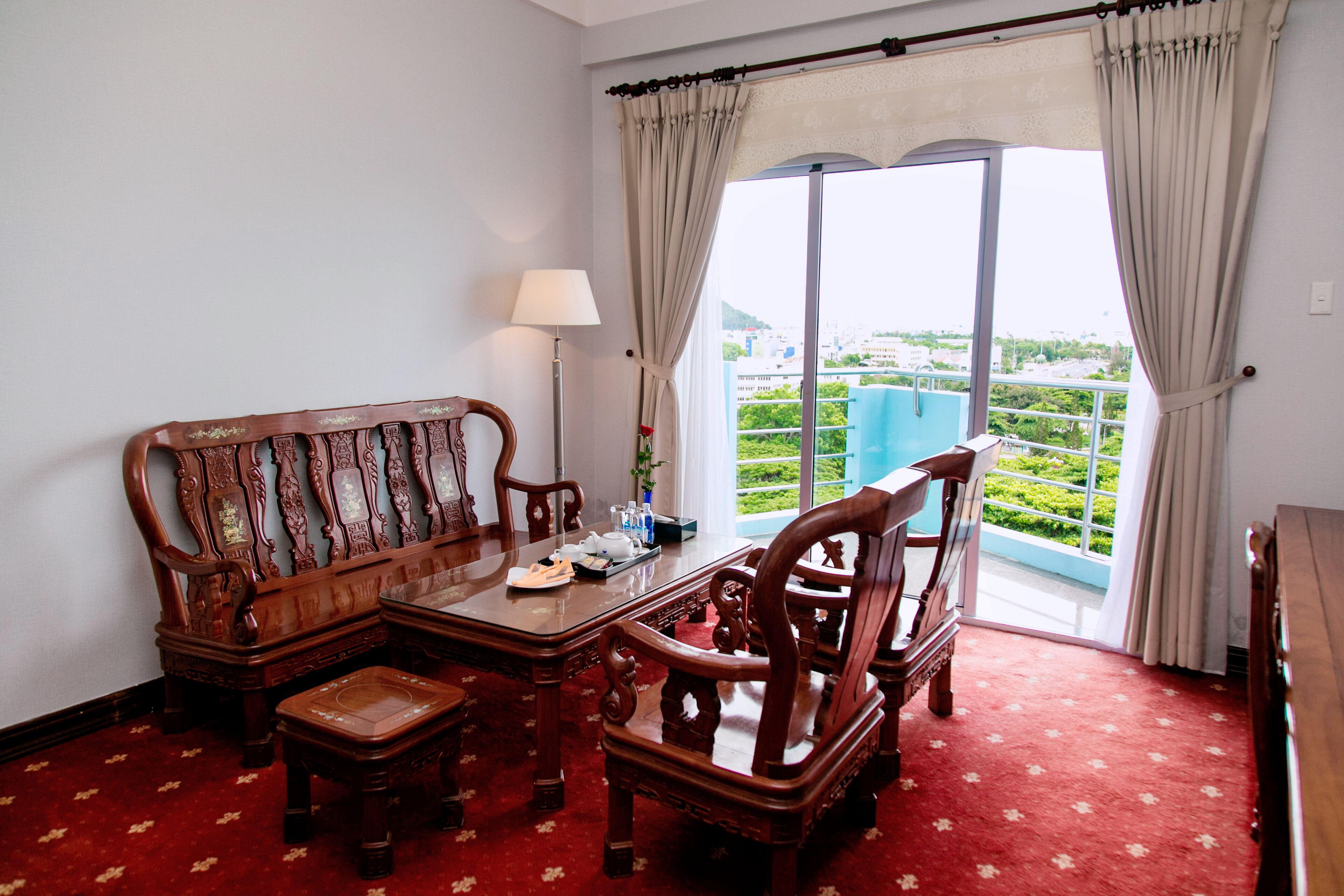 A traditional wooden sofa set with a matching armchair and ottoman, a small table, and a floor lamp. The room features a red carpet and a large window with curtains, offering a view of a green landscape.