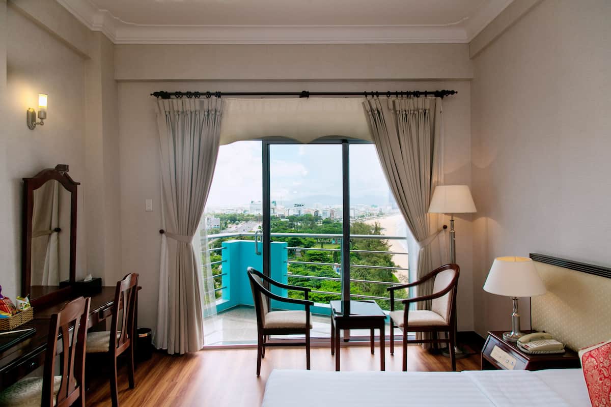 A hotel room with a large window, a bed, a desk with chairs, a lamp, and a view of greenery and buildings.