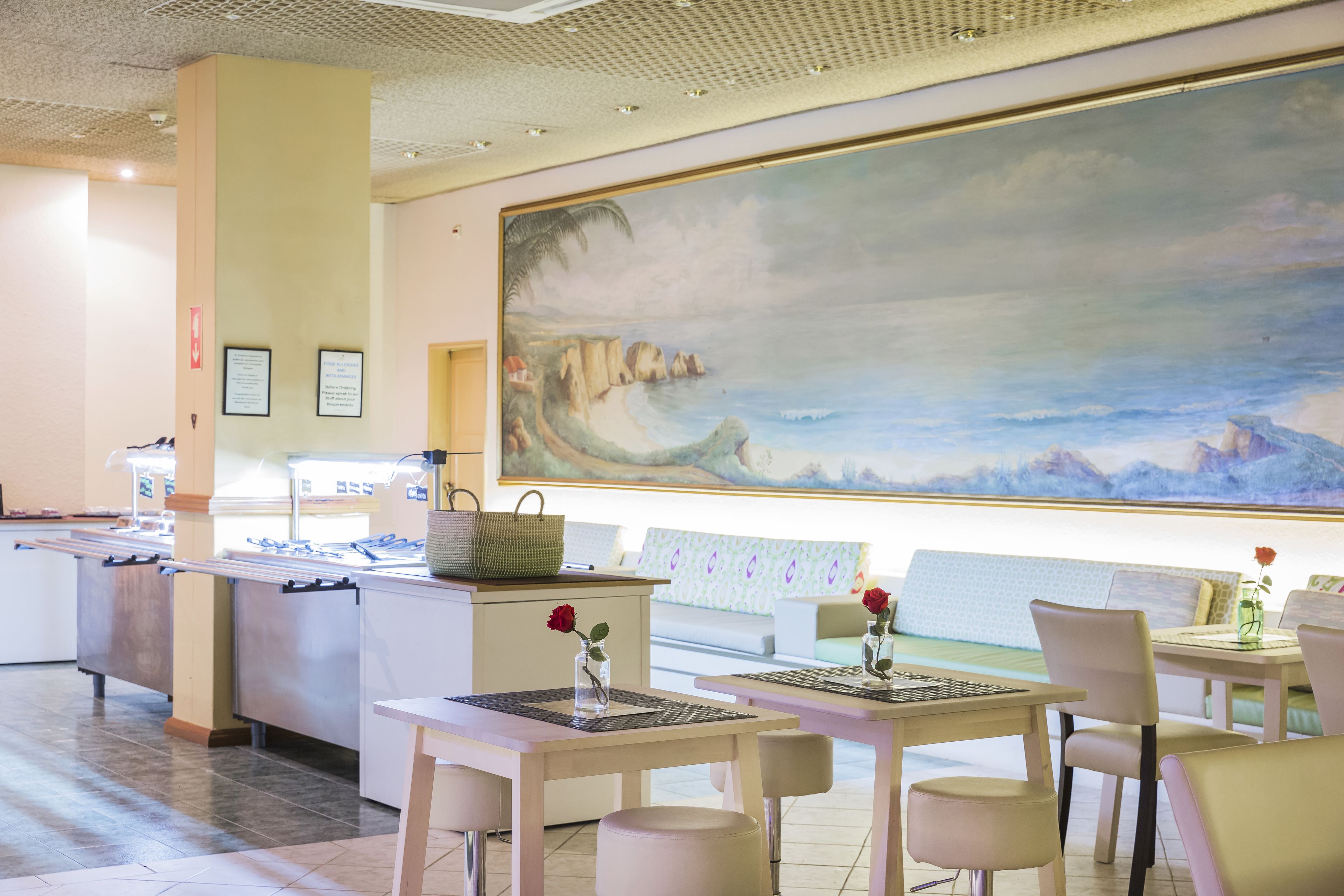 Een restaurant met een grote muurschildering van een kustlandschap, tafels met stoelen en een buffet.