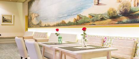 Un comedor con mesas y sillas blancas, un mural de un paisaje costero y una zona de estar con cojines estampados.
