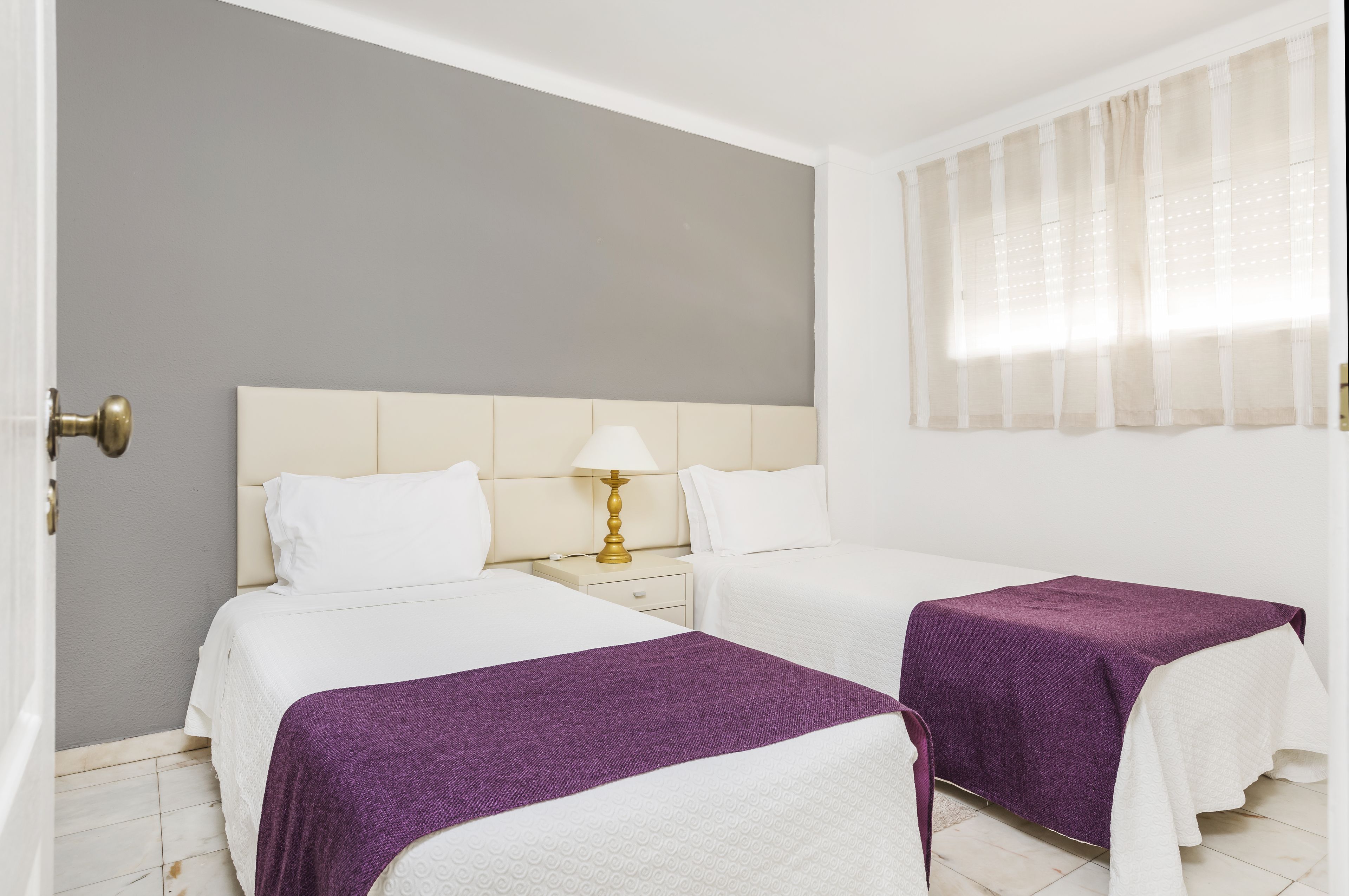 Habitación de hotel con dos camas, una mesita de noche con lámpara y una ventana con cortinas translúcidas.