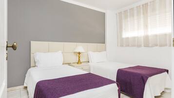 Habitación de hotel con dos camas, una mesita de noche con lámpara y una ventana con cortinas translúcidas.