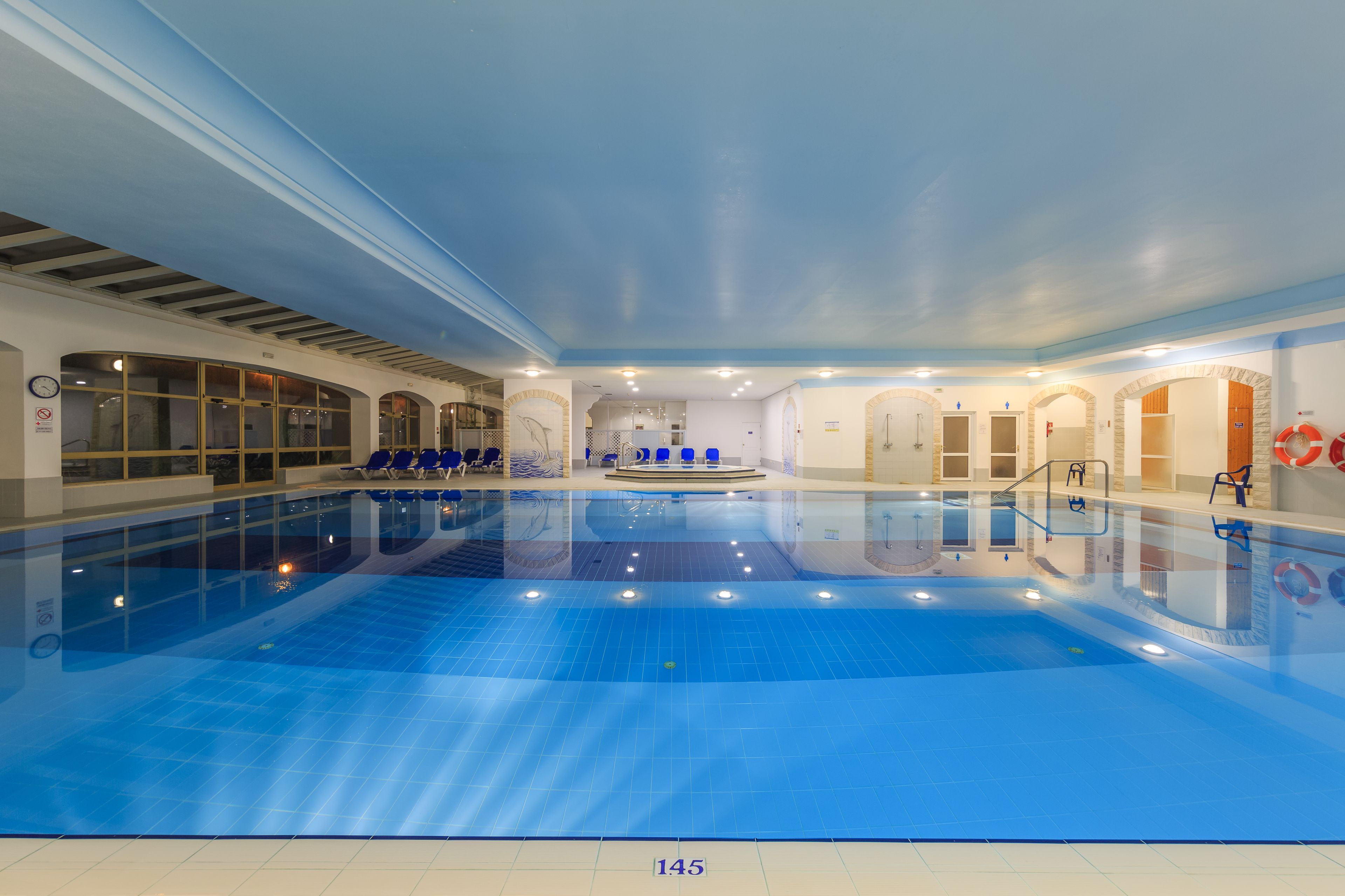 An indoor swimming pool with clear water, blue tiles, and a central area with blue chairs.