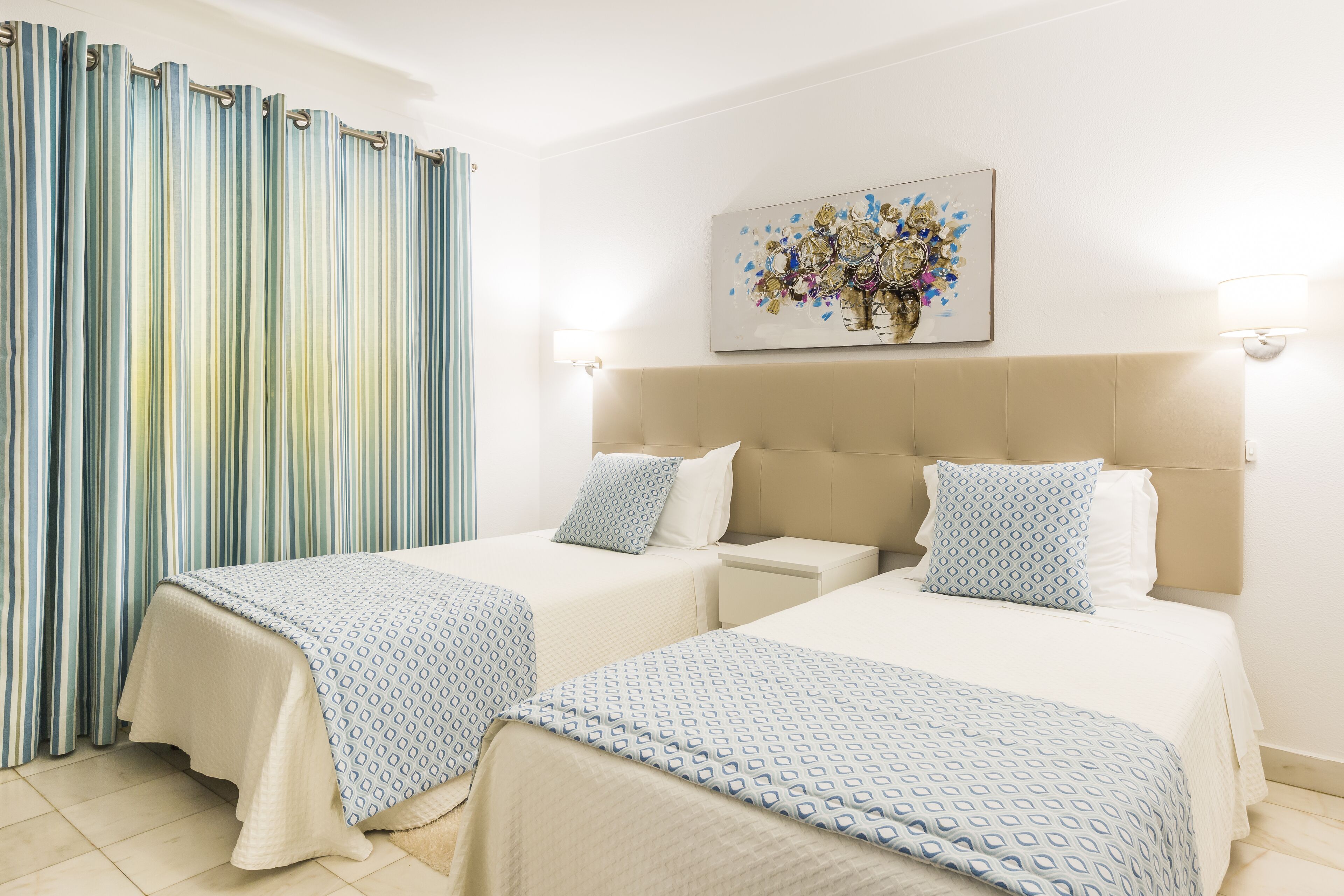 A hotel room with two beds, a striped curtain, a painting, and a nightstand.