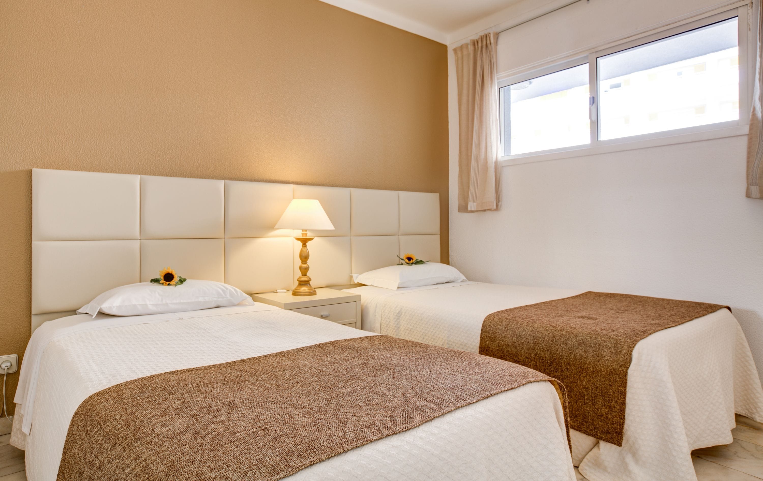 A hotel room with two beds, a nightstand with a lamp, and a window with curtains.