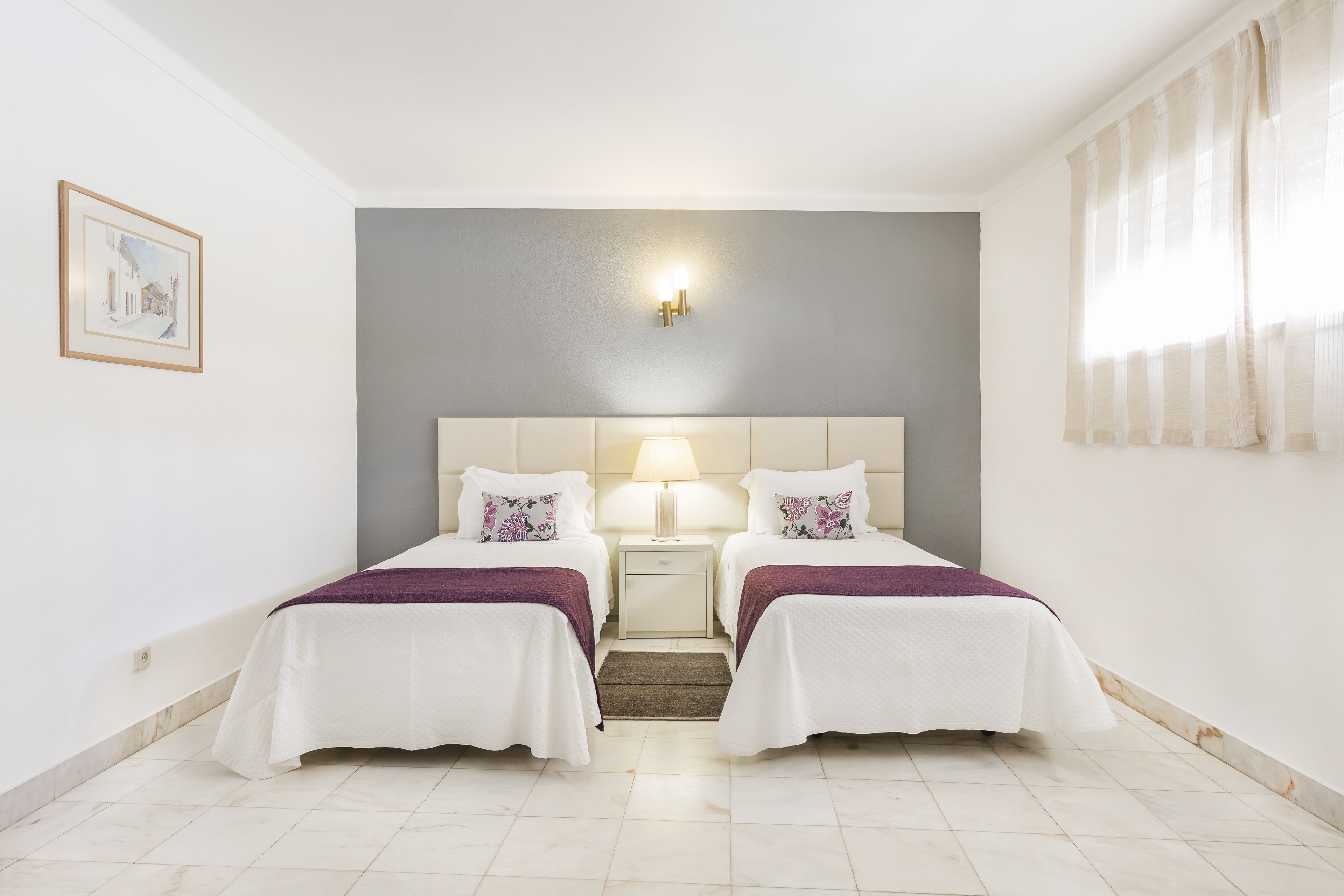 A hotel room with two beds, a nightstand with a lamp, a framed picture on the wall, and a window with curtains.