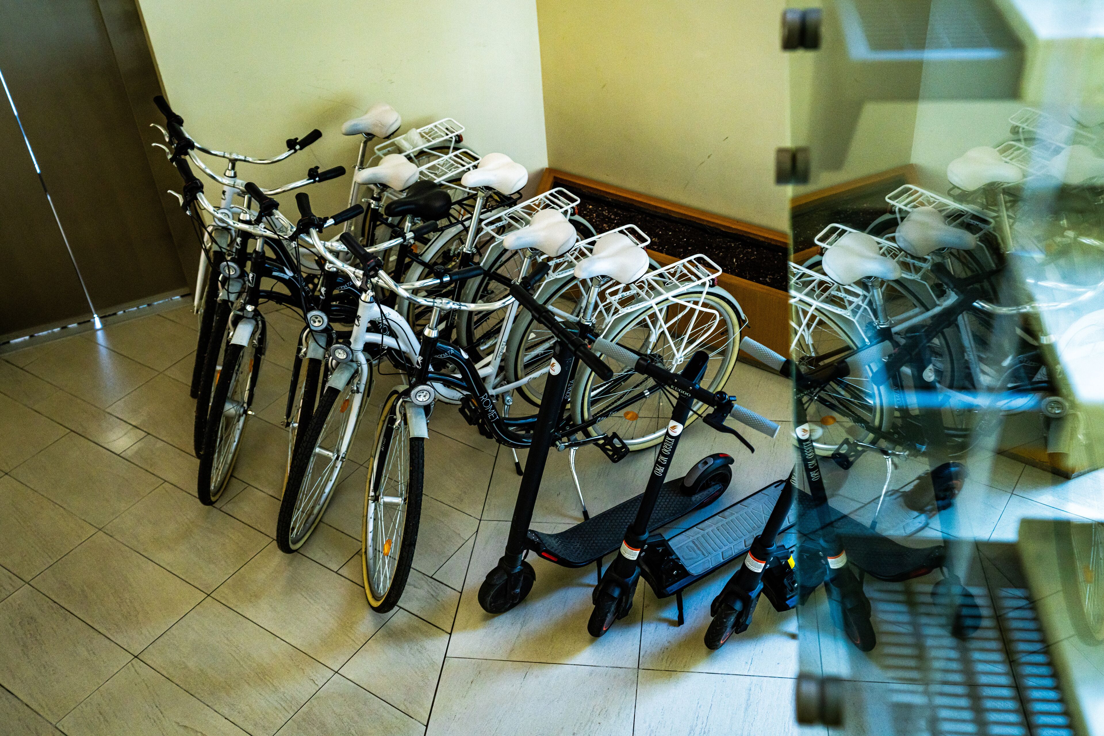 A row of bicycles with attached storage baskets, parked in a room with tiled flooring and a wooden floorboard border.