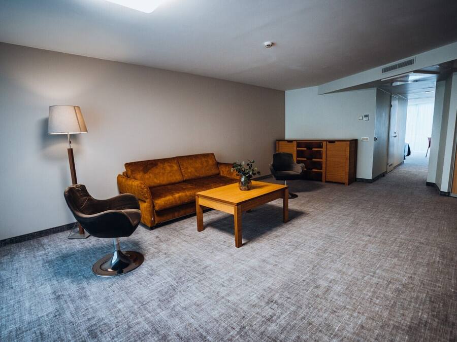 Un salon comprenant un canapé, un fauteuil, une table basse et une lampe. Une porte donne accès à une autre pièce.