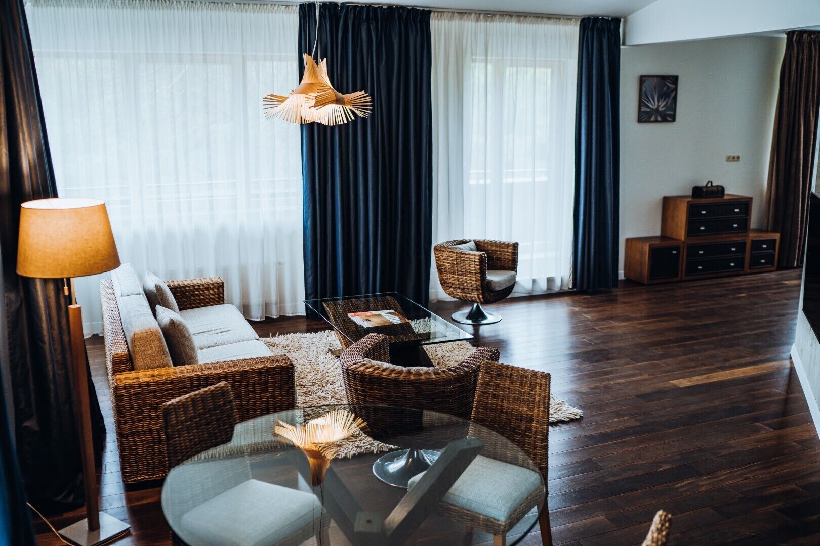 A modern living room with a sofa, two wicker chairs, a glass coffee table, and a hanging light fixture.